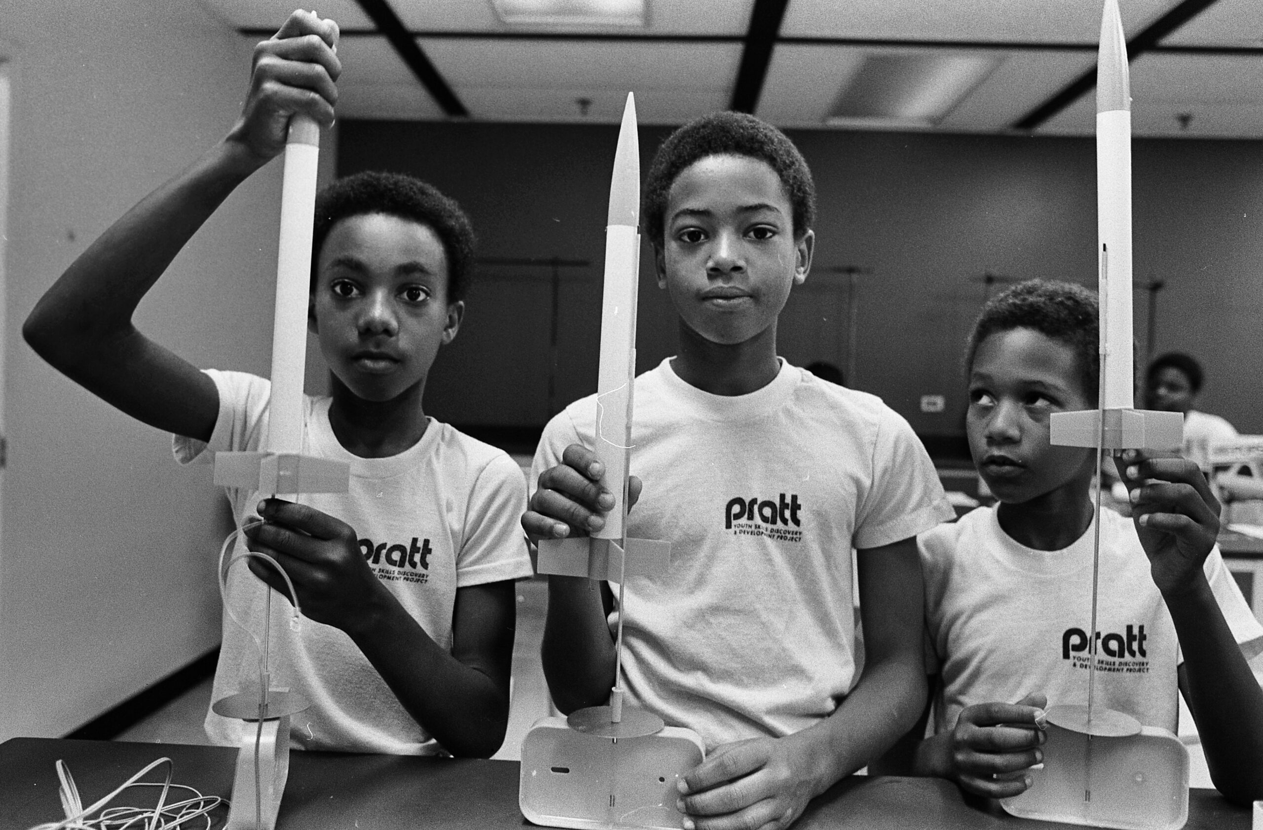 三个小男孩在教室里的黑白照片, 所有人的左胸前都印有“澳门足球博彩在线青年技能发现与发展项目”的字样, 在一张桌子上向照片的摄影师展示他们的火箭模型和配套的发射器.