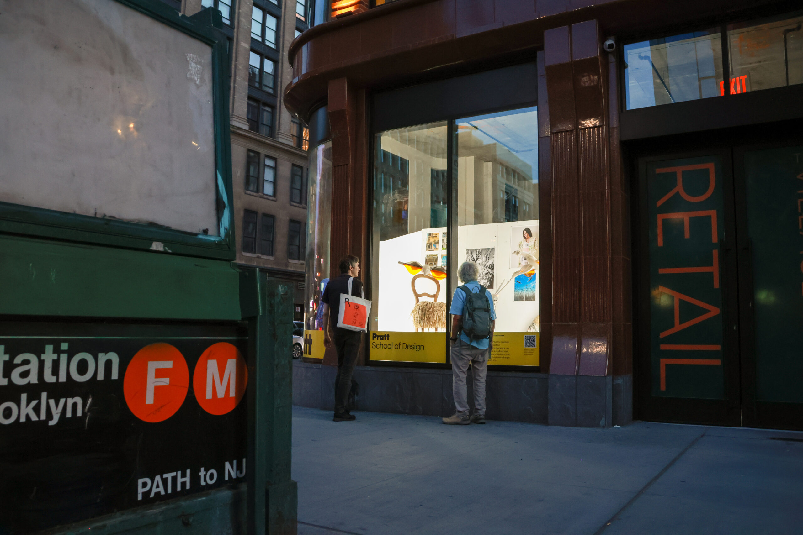 Two people stand outside a brightly lit storefront featuring a display for the Pratt School of Design. The window showcases various artworks, including a colorful sculpture and photographs. Nearby, a sign indicates a subway station with the "F" and "M" train symbols, and another sign labeled "PATH to NJ" is visible. The scene captures an urban evening atmosphere with buildings in the background.