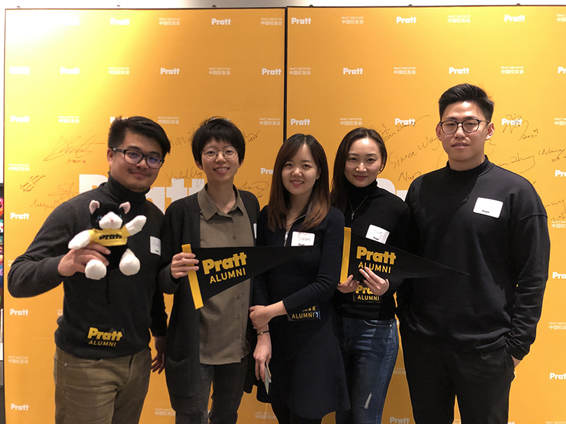 A group of five people standing in front of a yellow background with 'Pratt Alumni' written on it. They are smiling and holding black and yellow 'Pratt Alumni' pennants. The person on the far left is also holding a small stuffed animal. All participants are wearing name tags, dressed in casual or business casual attire, and the group is posed closely together.
