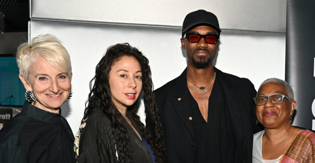 A group of four people standing side by side, smiling at the camera. From left to right: an older woman with short, platinum blonde hair, wearing a dark outfit; a woman with long dark wavy hair and a floral-printed jacket; a man with a goatee, wearing sunglasses, a black hat, and black clothing; and an older woman with short gray hair and glasses, dressed in a colorful shawl.