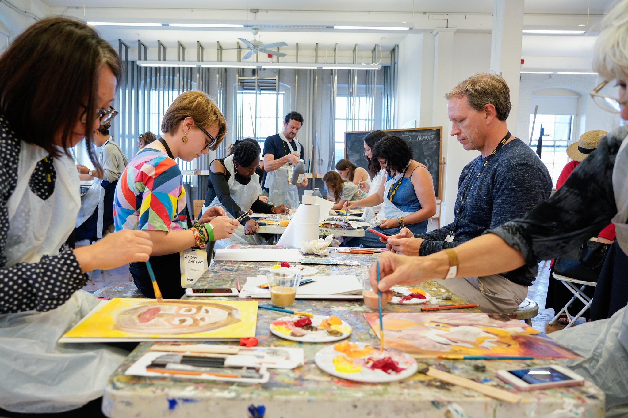 A group of adults seated around long tables in a bright classroom studio, actively painting and creating artwork. Each person wears a plastic apron and uses paintbrushes, palettes, and paper or canvas. The foreground shows colorful portraits and palettes with red, yellow, and pink paint. The room is filled with natural light from large windows, and a chalkboard is visible in the background.