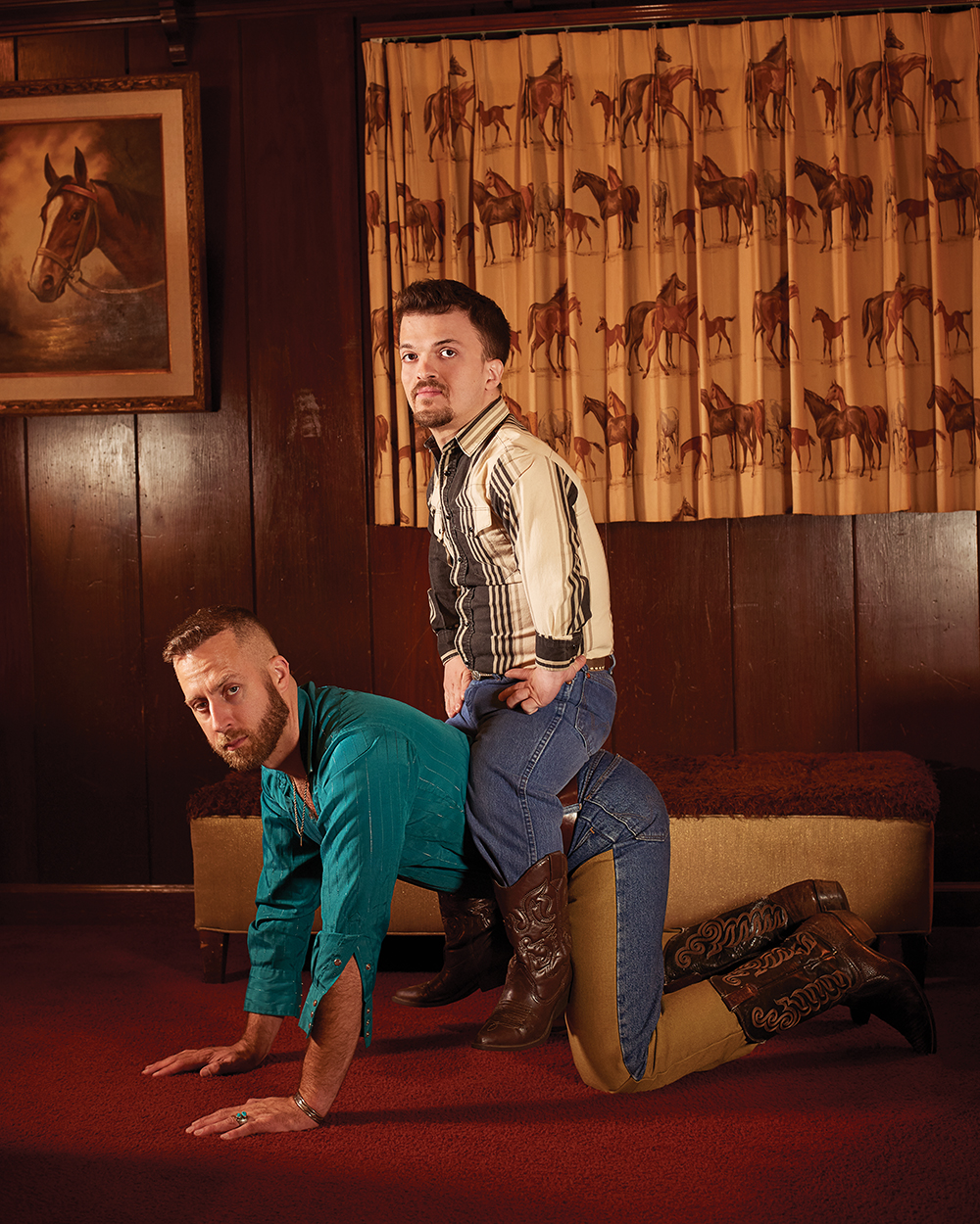 A man with dwarfism rides the back of a man of average stature. They wear western-themed clothing and are in a wood-paneled room decorated with a western theme, including curtains printed with horse illustrations and a painting of a horse. The figures look directly at the viewer with forthright and neutral facial expressions.