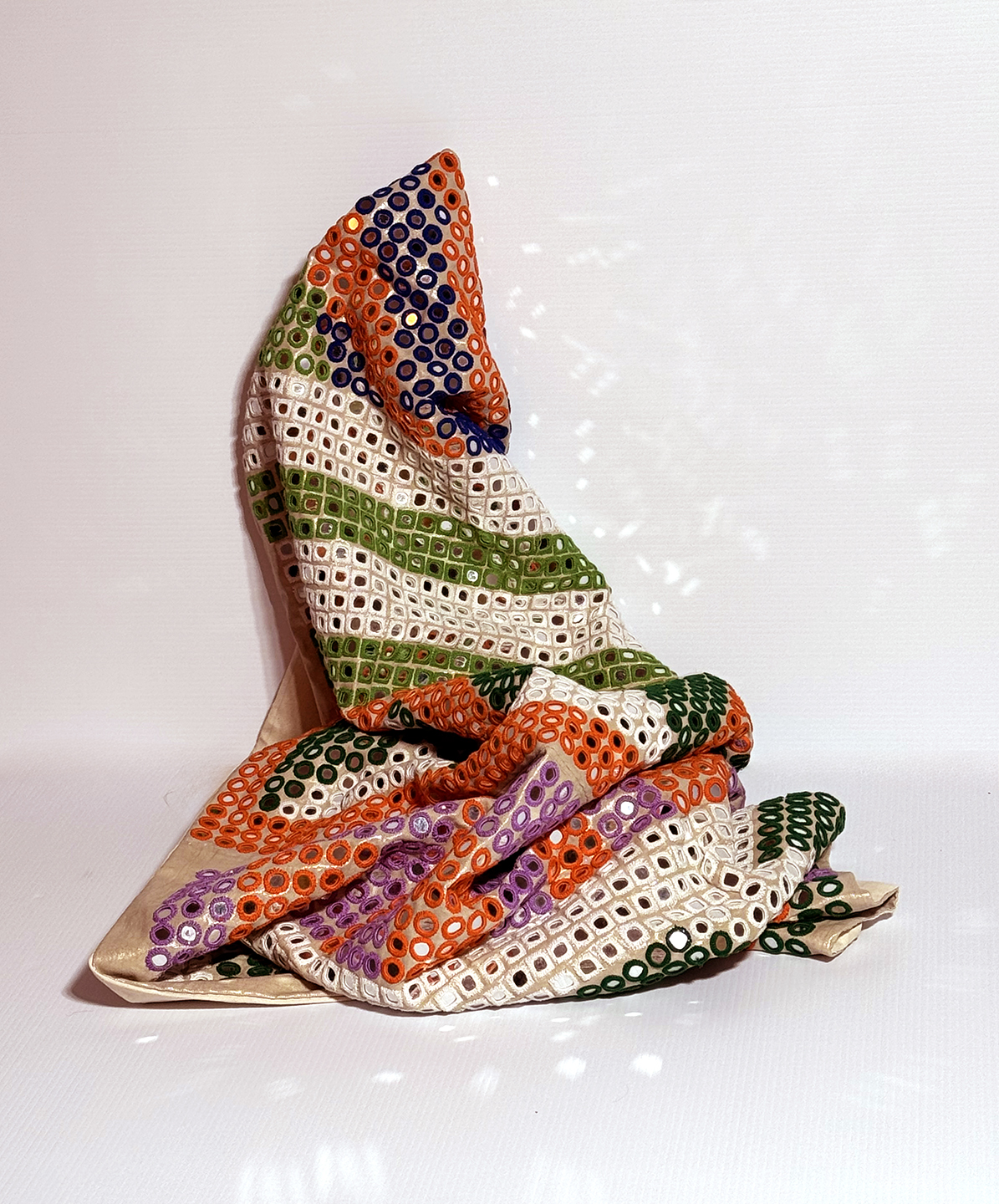 A work resembling a colorful blanket with many grommets piled against a wall.