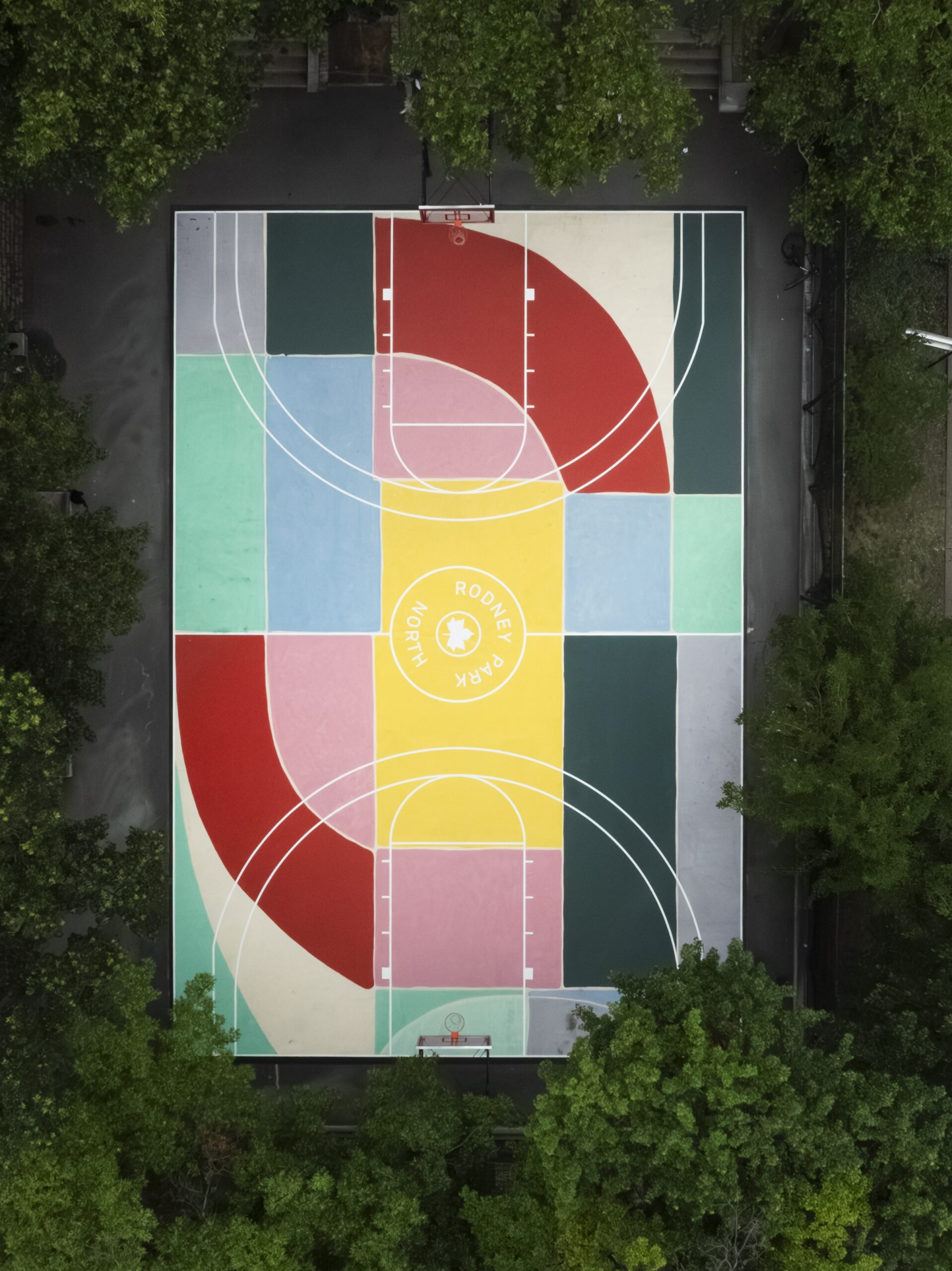 Photographed from above, a colorful basketball court with segments painted forest green, teal, powder blue, red, grey, and taupe. At the center of the court, in a vibrant yellow square, is the logo of Rodney Park North. The court is surrounded by leafy green trees.