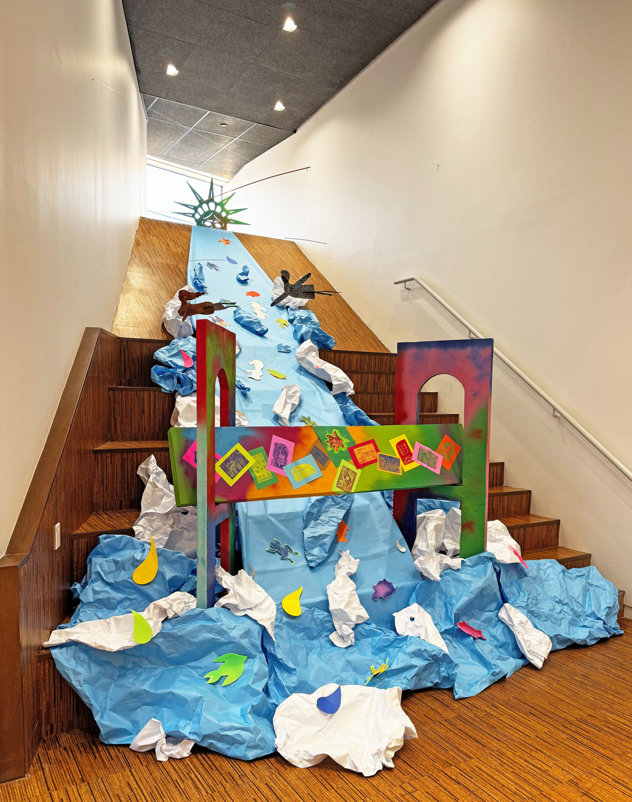 An art installation. Crumpled blue and white construction paper forms a waterfall down a wooden staircase. In the waterfall are colorful cut-outs of birds, frogs, and abstract forms. At the top of the waterfall is a cut-off of the statue of liberty's head, with only her crown and the top half of her face peering over the top of the waterfall. At the base of the waterfall is a colorful wooden bridge with designs pasted to the main span. Two cutouts of human figures appear to soar above the waterfall, their arms and legs outstretched.