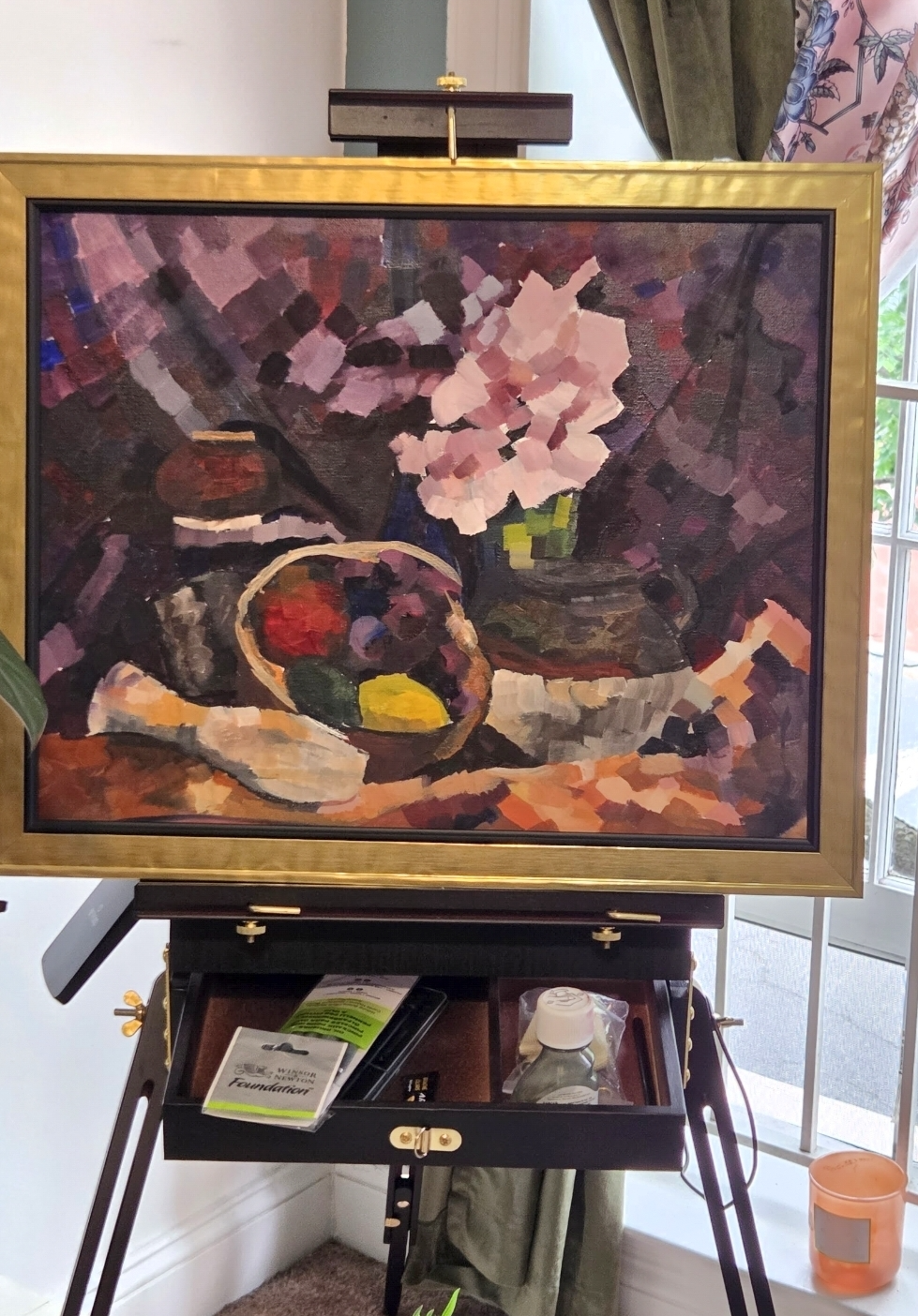 A still life painting of a vase of flowers and fruit basket in an impressionistic style in purple and ochre tones in a gold frame on an easel.