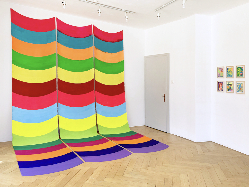 In a gallery, a large-scale installation is hung on the walls and drapes onto the floor. The installation is composed of three pieces of fabric, hung side by side, that together form colorful wavy stripes in vibrant, saturated hues of cerise pink, teal, orange, lime green, butter yellow, powder blue, chartreuse, lime green, forest green, magenta, indigo, and violet. On an adjacent wall is installed a 3 x 2 grid of small works depicting abstract shapes and patterns in similarly vibrant hues.