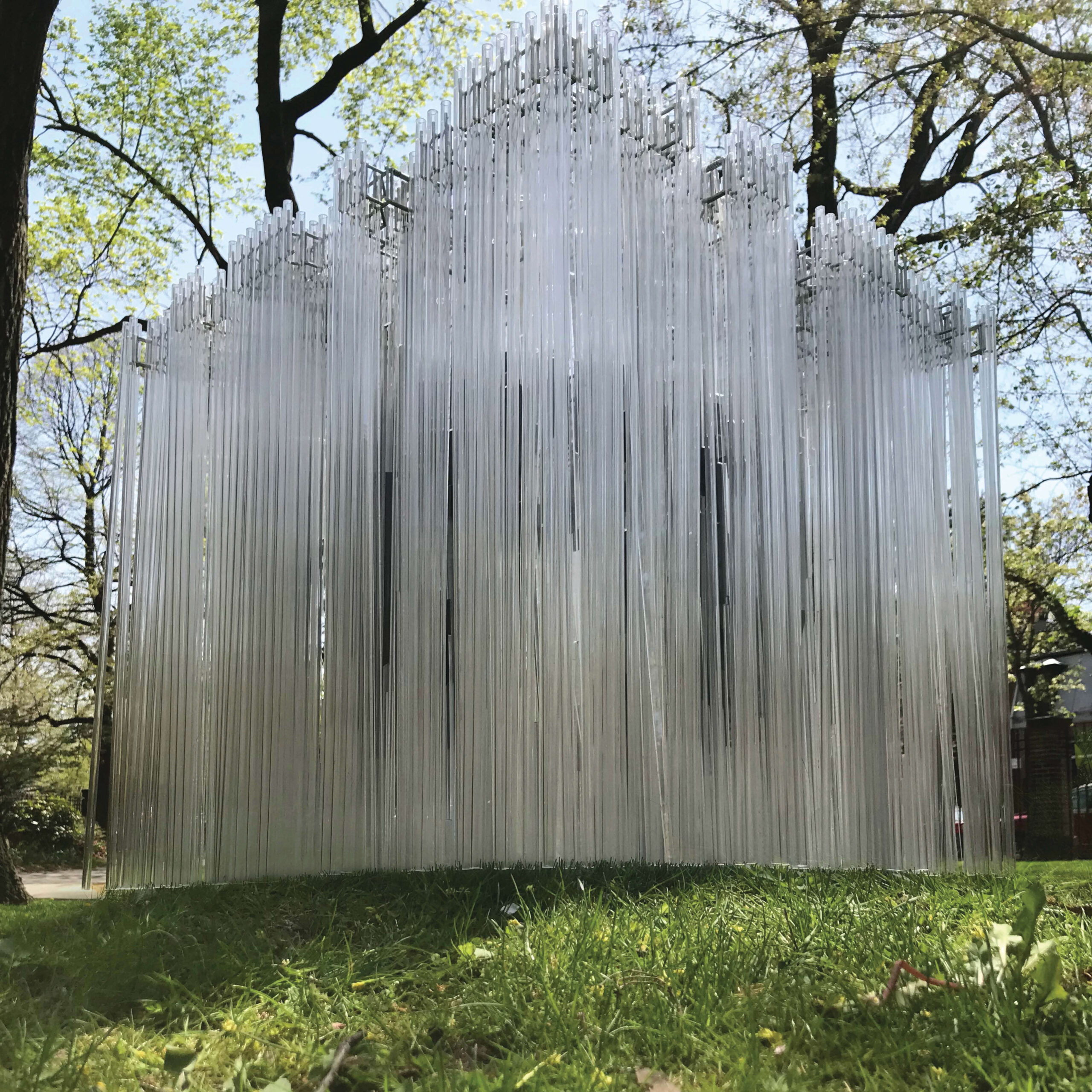 A cubical structure made out of tall glass filaments stands atop grass with trees in the background.