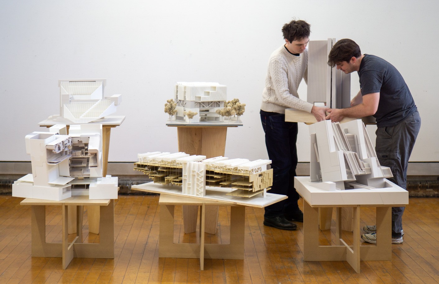 Two students stand in a studio space adjusting a large white architectural model on a table. Several detailed architectural scale models are displayed on wooden pedestal tables arranged across a hardwood floor. The models include cutaway sections, layered structural elements, and small landscape features. The setting appears to be an architecture studio or exhibition space with a plain white wall backdrop.