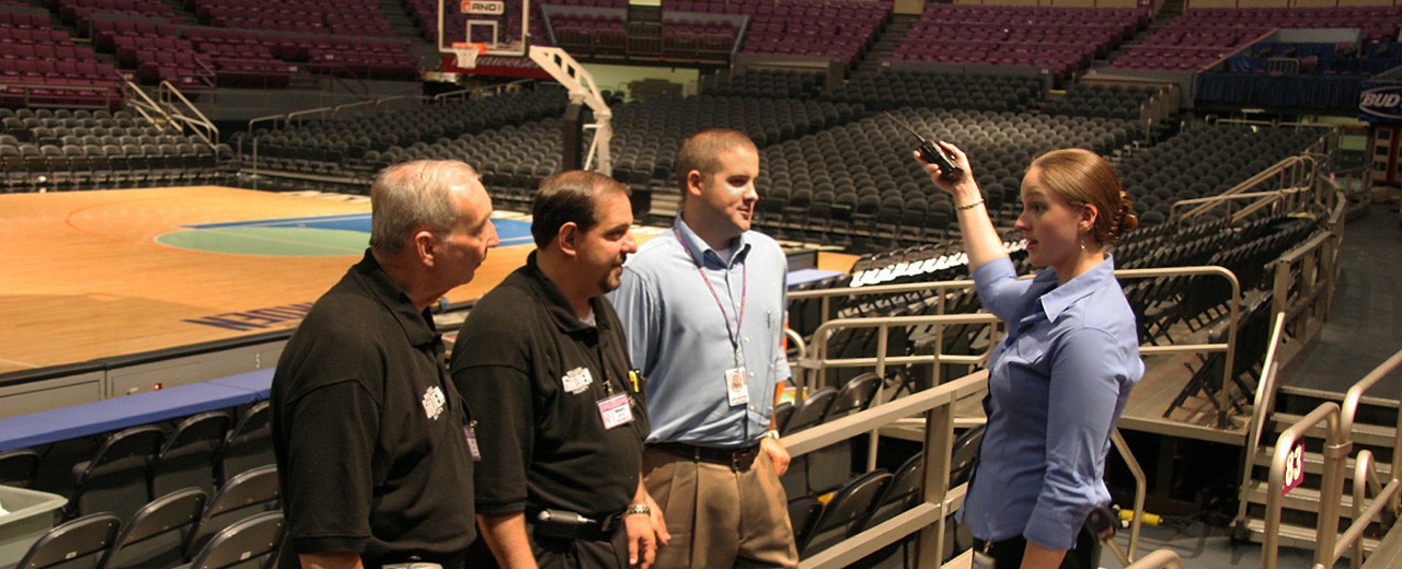 A group of people listen to a young woman giving instructions while holding a handheld radio. They are standing on the bleachers of a large stadium.