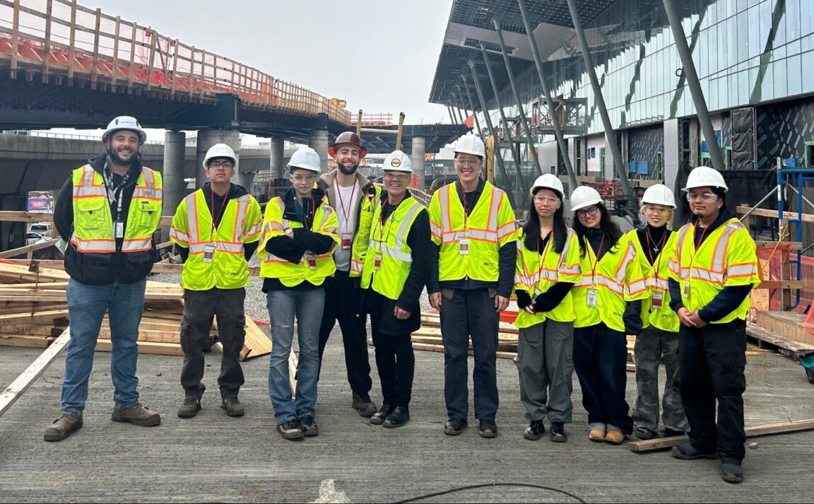 Group of students and instructors wearing high-visibility safety vests and white hard hats standing together at an active construction site. Construction materials, scaffolding, and a large building structure are visible behind them, indicating a site visit or field experience related to construction management.