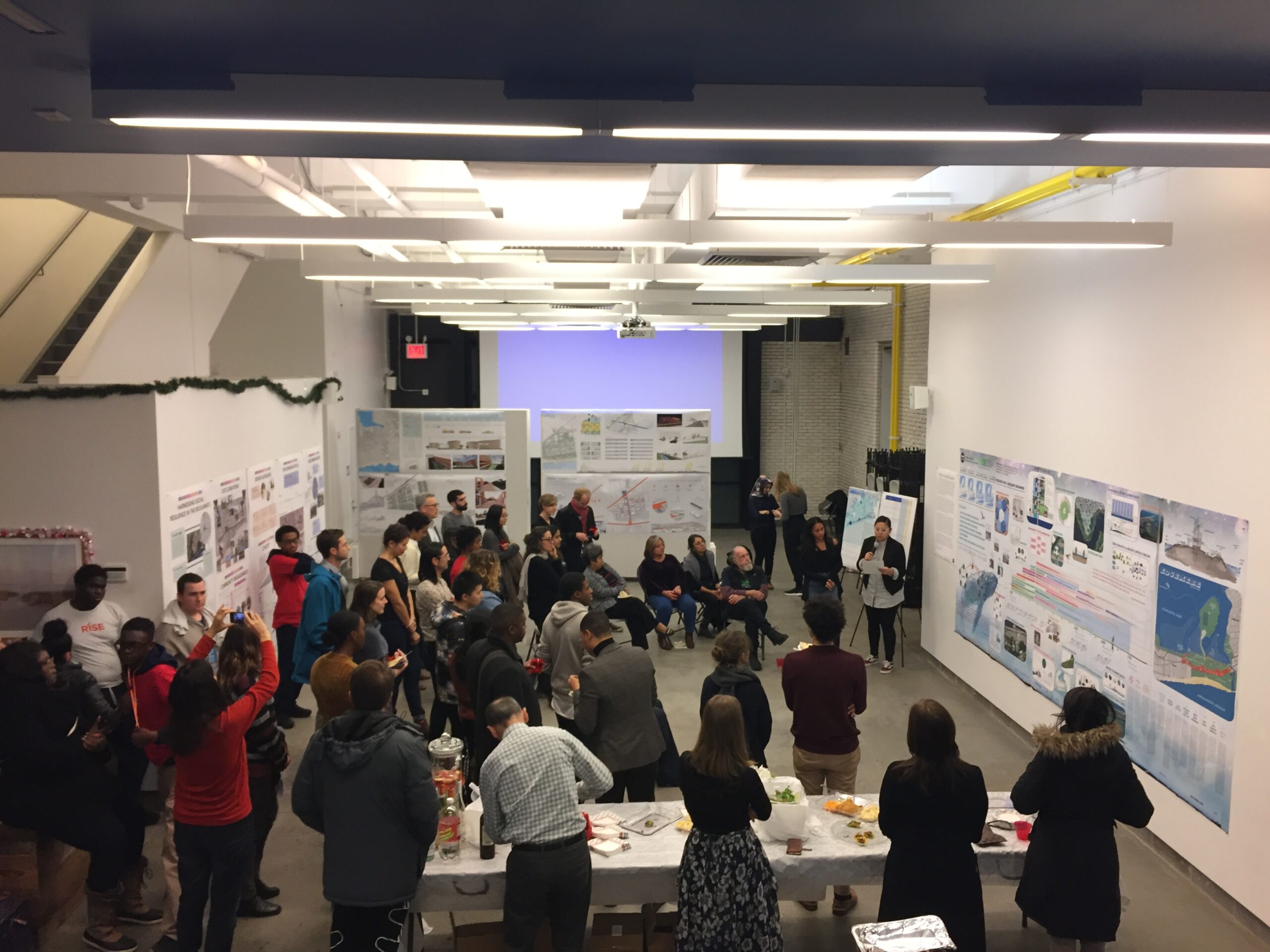 A group of people in a large room. Designs are pinned to the wall and someone is presenting to the group.