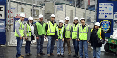 A group of students pose for a picture with a construction site in the background. They are wearing construction site gear, including white helmets and yellow safety vests.