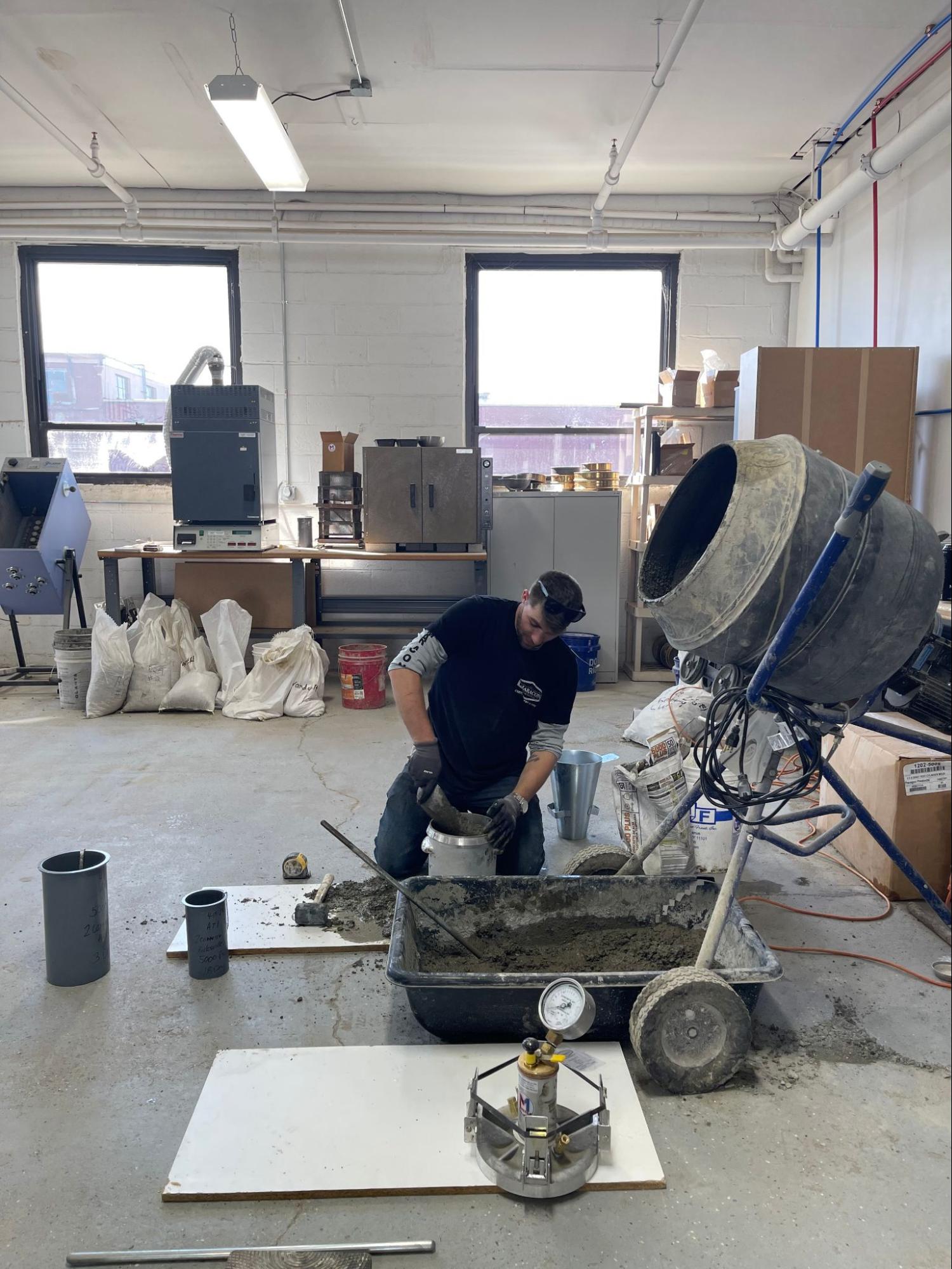 A person works in a materials lab, mixing concrete from a large batch mixer into testing molds. Various lab equipment, bags of materials, and industrial machines are visible in the background.