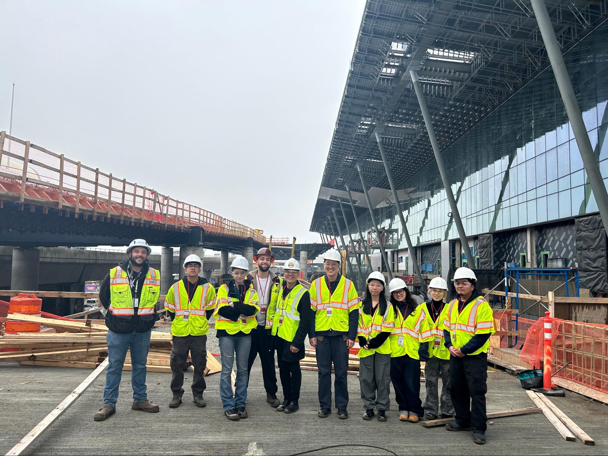 A group of people in hard hats and high-visibility safety vests stand together at an active construction site in front of a large modern glass building with exposed steel framing. Wooden scaffolding, orange barriers, and construction materials surround them.