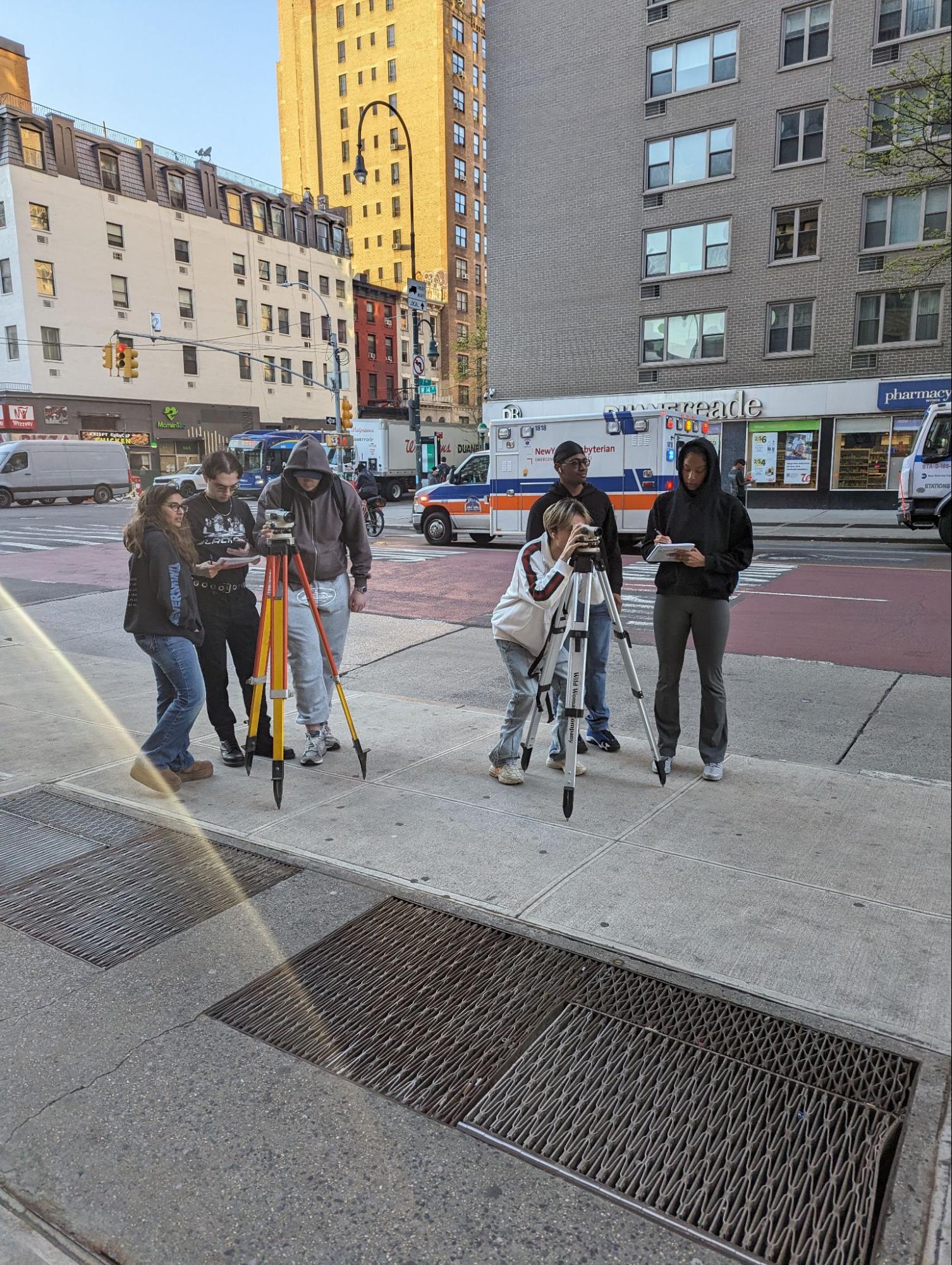 A group of students on a city sidewalk use surveying equipment with tripods near a busy intersection lined with tall buildings, vehicles, and a passing ambulance. They appear to be conducting field measurements or urban planning exercises.