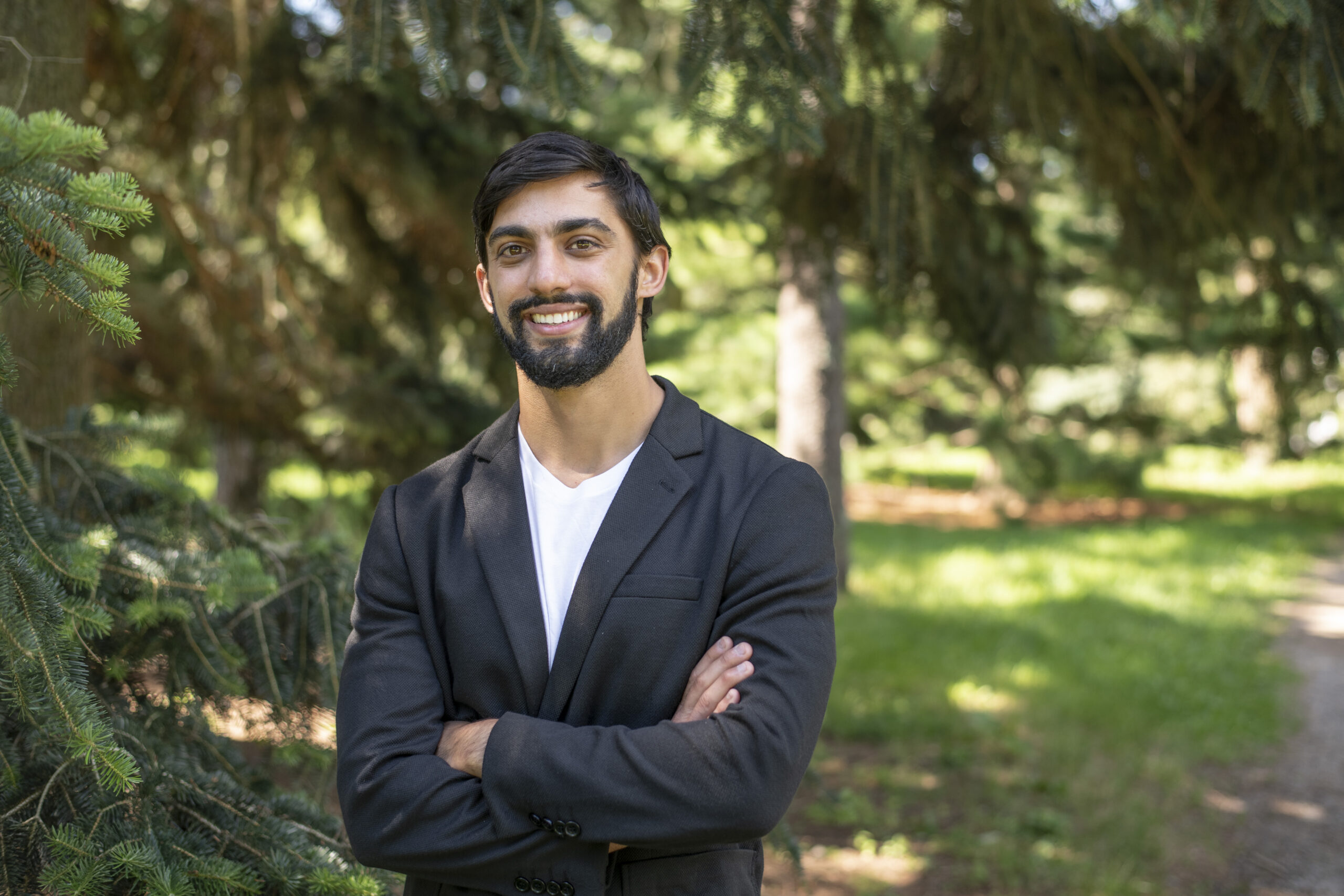 headshot of Jacob Suissa, standing in a park, arms crossed, with pine trees behind him, and a path bordering with a gravel road