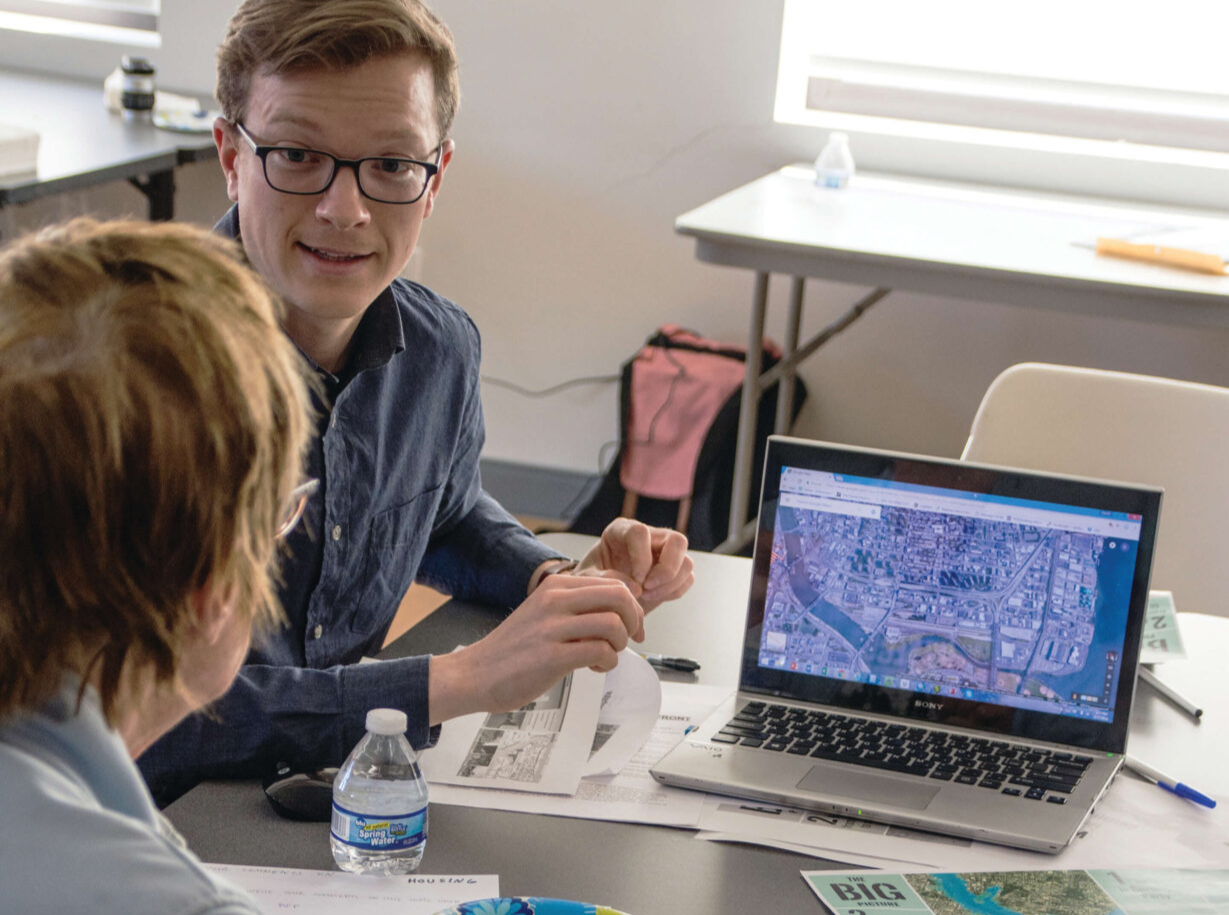 Two people discuss while sitting next to a table covered in documents and blueprints. There is a computer with a map on its screen resting on the table.