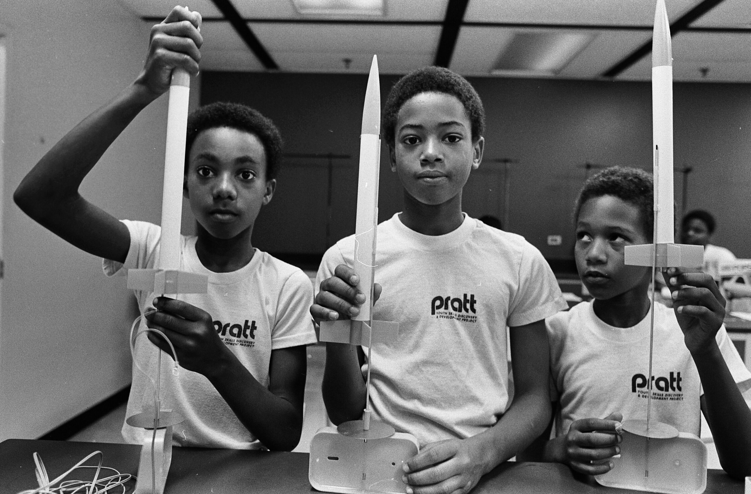 A black and white photograph of three young boys in a classroom, all wearing shirts with the text “PRATT YOUTH SKILLS DISCOVERY AND DEVELOPMENT PROJECT” printed on the left chest, displaying their model rockets and accompanying launchers to the picture's photographer on a table.