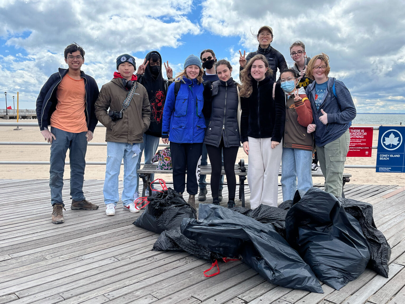 A group of people posing for the photo on the deck of the Coney Island Beach. There are a few trash bags in front of them, possibly implying that they were cleaning up the beach.