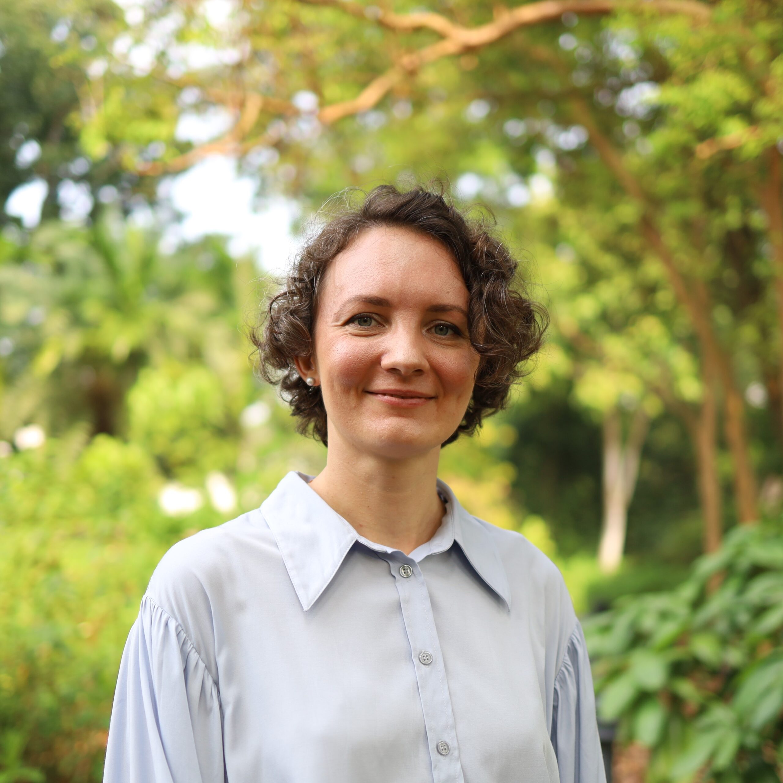 photo of Yulia Dzyuban, standing in forest, with foilage and trees in background, subject in foreground, smiling, wearing collared dress shirt