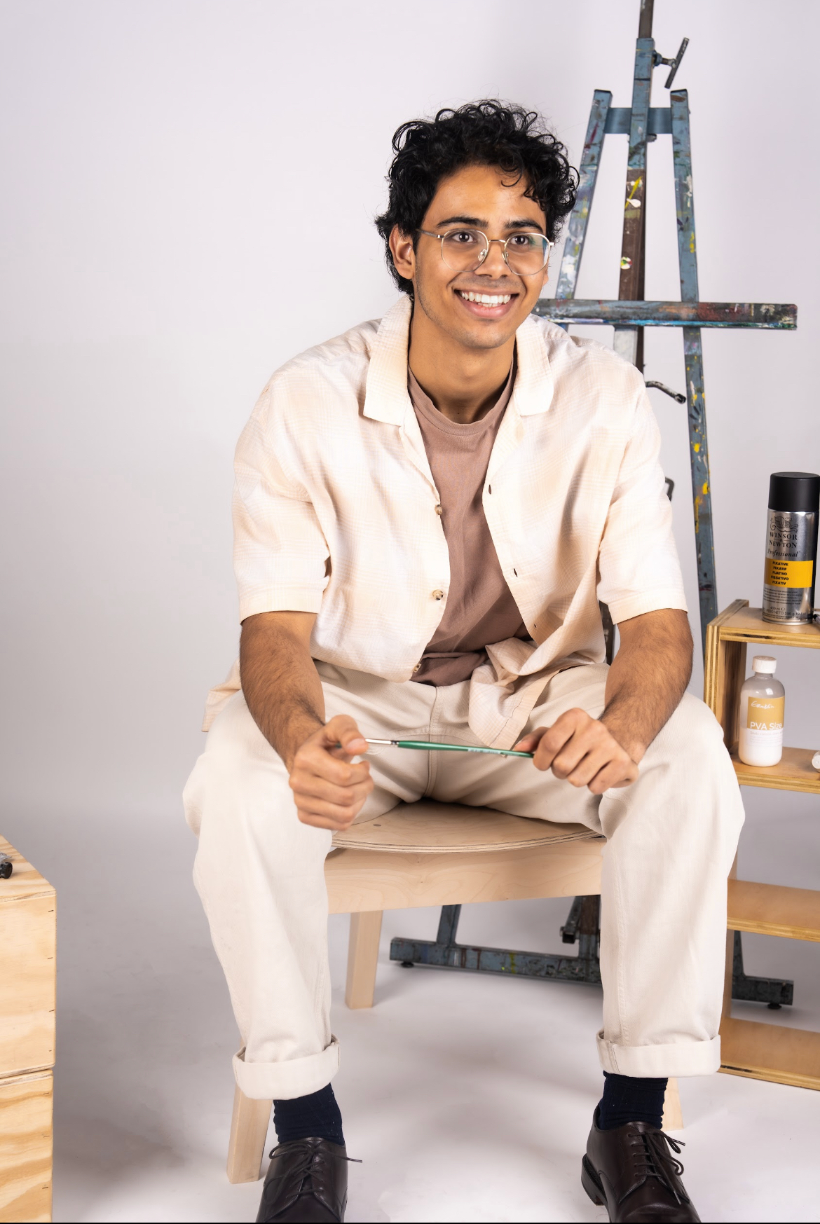 A young man with curly black hair, glasses, and a warm smile sits on a wooden stool in an art studio. He is wearing a beige short-sleeve button-up shirt over a brown t-shirt, cream-colored pants, and dark leather shoes. He holds a paintbrush in both hands while looking slightly off-camera. Behind him, a paint-splattered easel leans against the wall, and a small wooden shelf holds art supplies, including a can of spray paint and a bottle of PVA size. The background is a clean, neutral-toned studio setting.