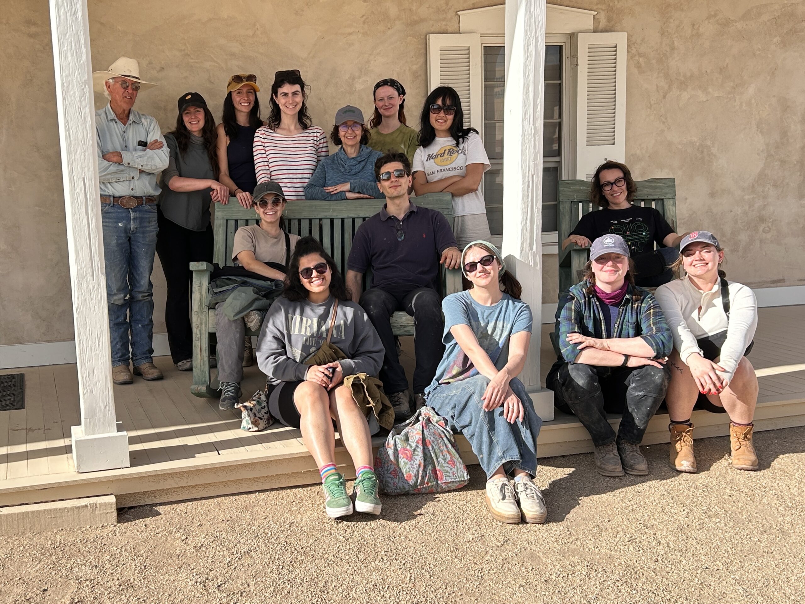 A group of thirteen people, including students and an older man in a cowboy hat, pose together on the porch of a stucco building. Some sit on green benches while others kneel or sit on the porch steps, smiling in the sun. Everyone appears casually dressed in work clothes, suggesting they have been participating in a hands-on outdoor project or workshop.