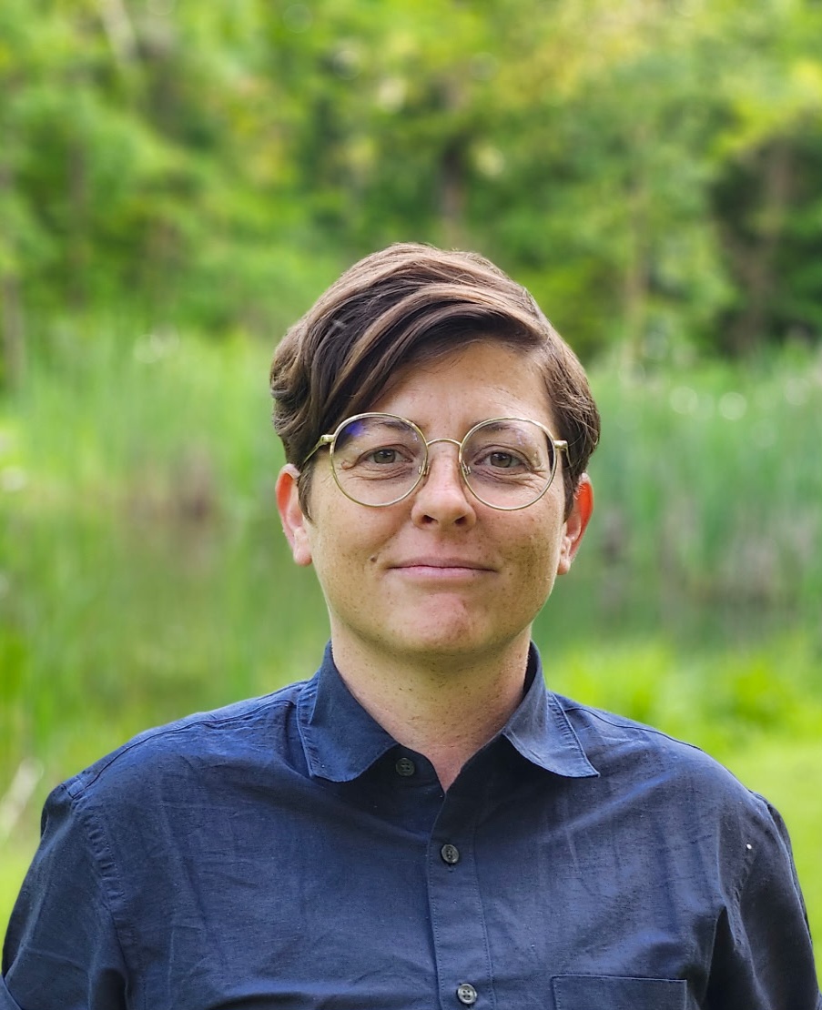 A person with short, dark hair and glasses stands outdoors in front of a blurred background of green foliage and tall grass. They are wearing a dark blue button-up shirt and looking directly at the camera with a calm, neutral expression. The natural light and setting give the photo a warm, serene feel.