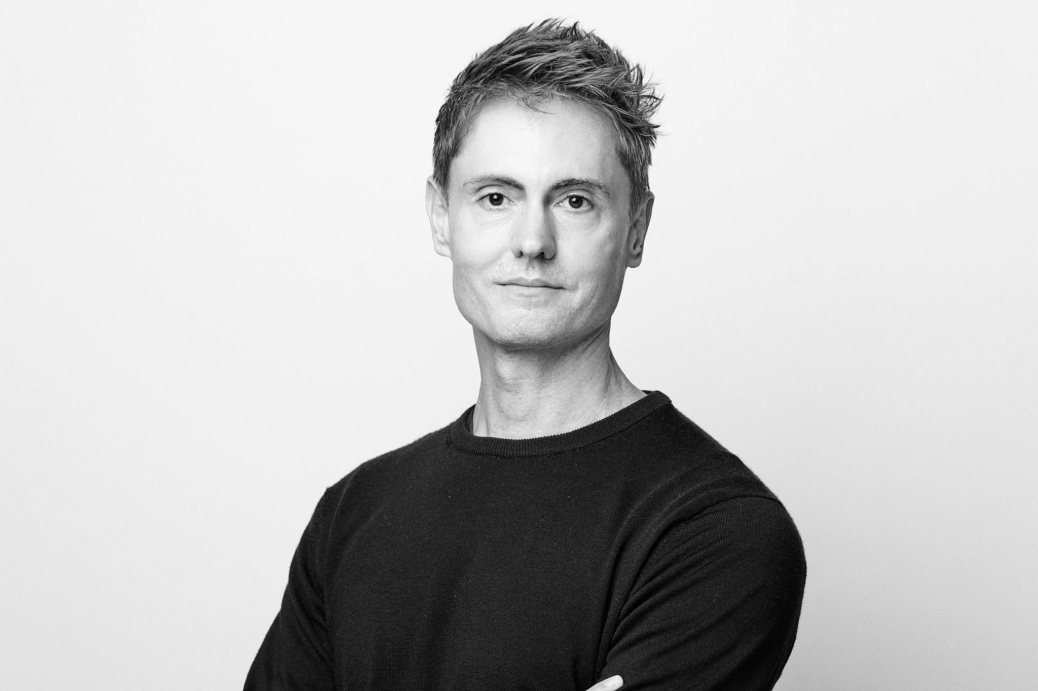 Black-and-white studio portrait of a man with short, textured hair wearing a dark crewneck sweater. He faces the camera with a neutral expression, arms folded, against a plain white background.