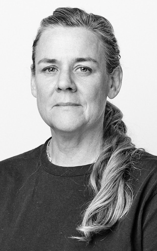 Black-and-white portrait of a woman with her hair pulled back in a low ponytail, wearing a simple dark top. She looks directly at the camera with a serious, steady expression, set against a plain white background.
