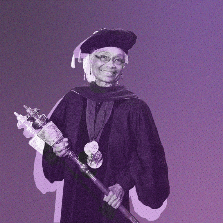 A woman in a graduation gown and cap smiles while holding a ceremonial staff adorned with ornate designs. She wears several medals around her neck and stands against a purple background, giving a sense of celebration and achievement.