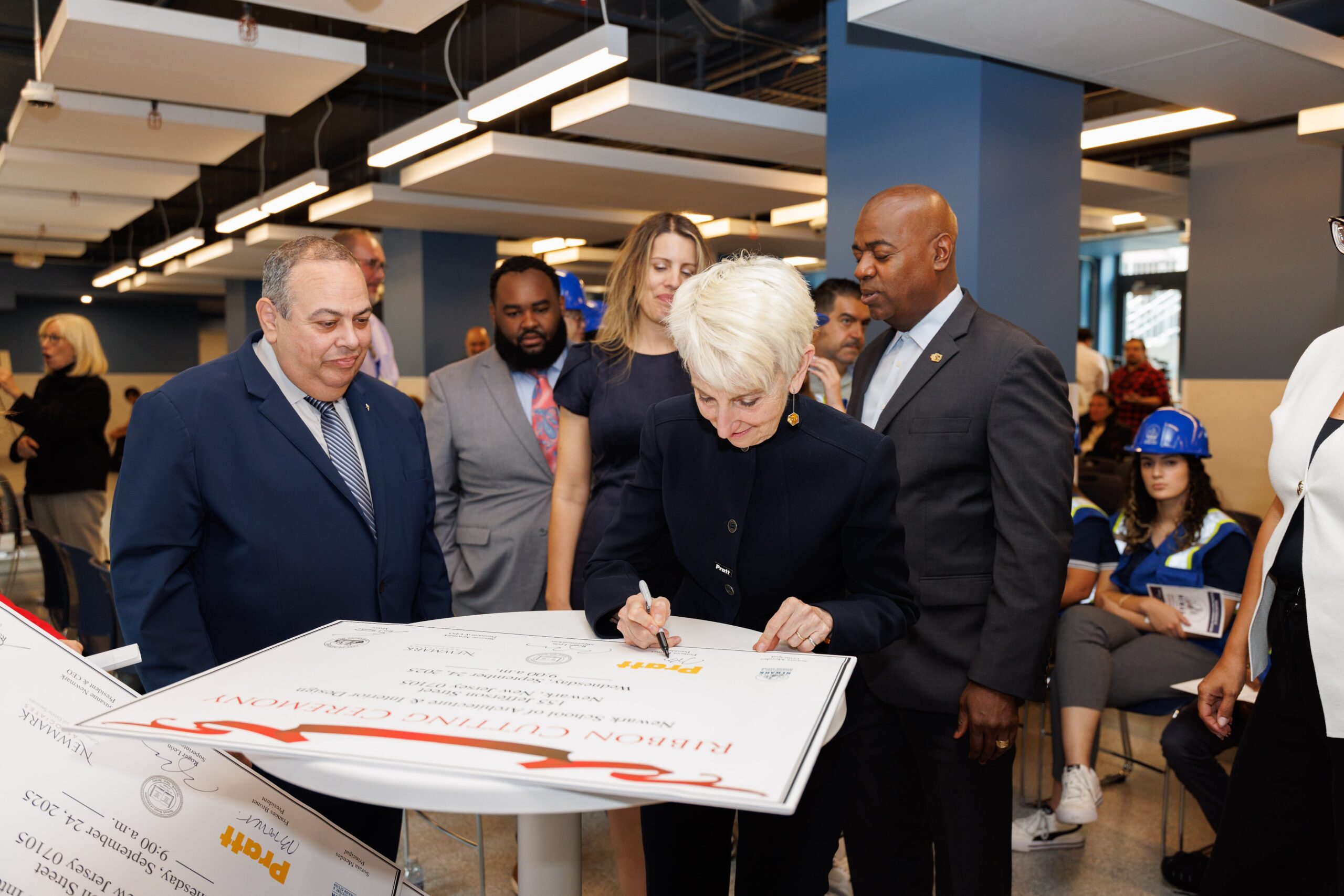 A group of people gathers for a ceremonial signing event in a modern indoor space. In the foreground, a woman with short white hair, dressed in a dark outfit, is signing a large poster on a table. Surrounding her are four men in suits and a woman in a blue dress, observing the signing. Some attendees in the background are seated, with a few wearing blue hard hats.