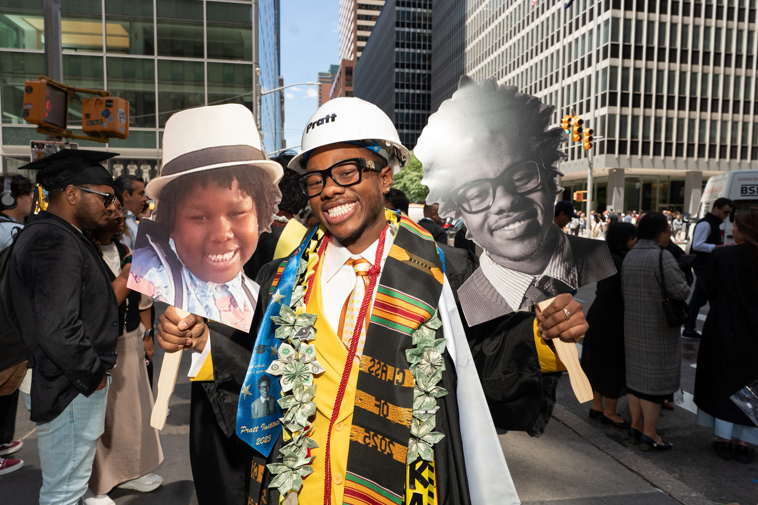 A smiling graduate in a black cap and gown, wearing a white hard hat, holds up two large cutouts of faces. One face is of a child with curly hair wearing a hat, and the other is of a man in glasses with styled hair. The graduate is adorned with a colorful stole featuring various patterns and pendant-like decorations. The background shows a city street with people and tall buildings.