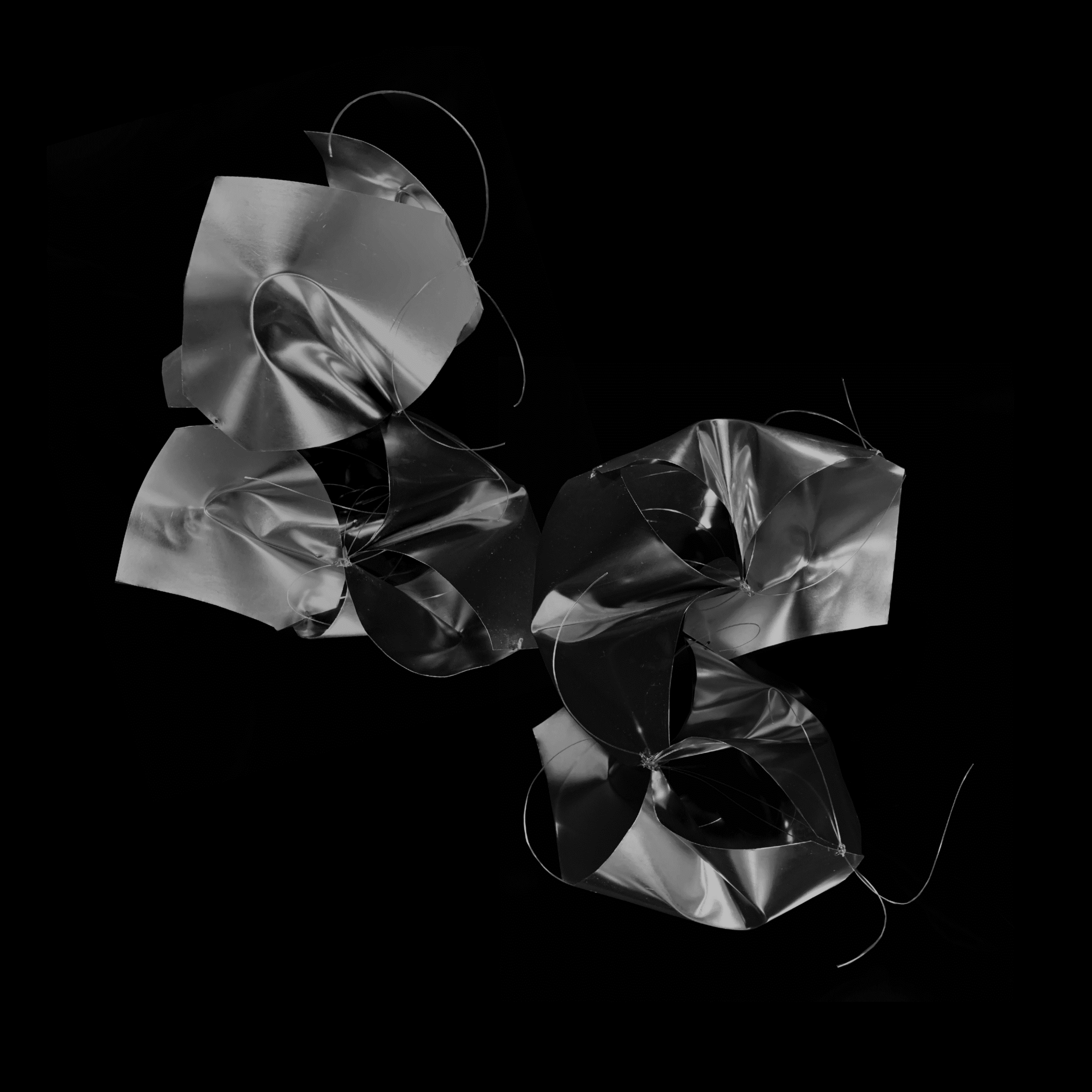 Three abstract shapes made of shiny, metallic-looking material are arranged against a black background. The forms appear crumpled and intertwined, with some thin wires protruding. The lighting accentuates the shiny surfaces, creating a sense of depth and movement.