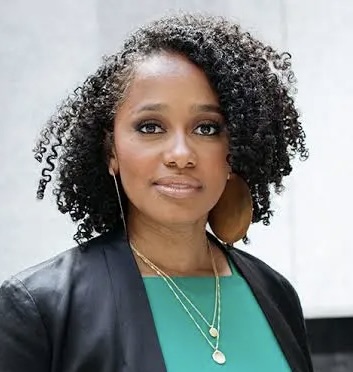 A professional headshot of a woman with curly hair wearing a black blazer over a green blouse, accessorized with layered gold necklaces and large earrings. She is standing in front of a light-colored urban background, looking directly at the camera with a calm, confident expression.