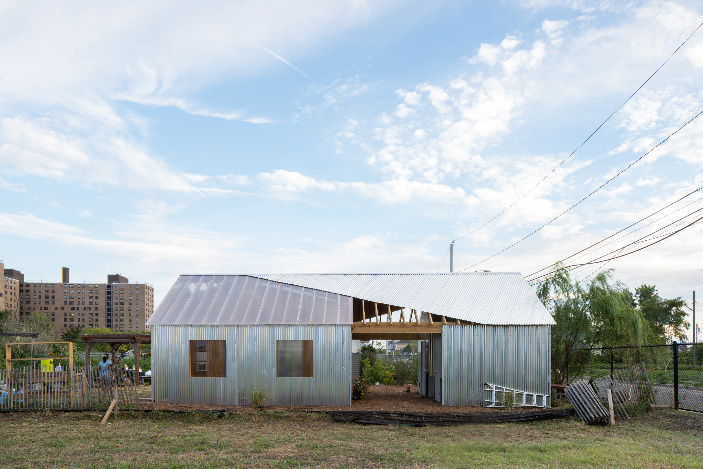 A modern, asymmetrical building made of corrugated metal and transparent roofing panels is depicted, surrounded by an open grassy area. In the background, tall apartment buildings are visible against a blue sky with wispy clouds. Electric wires stretch above the building, and a few trees and garden structures are situated nearby. A person can be seen in the foreground, tending to the garden.