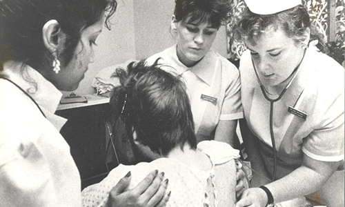 Black-and-white archival photograph showing three nurses providing medical care to a patient seated with their back to the camera. Two nurses in uniform use a stethoscope and attend to the patient while another supports them by placing a hand on their shoulder. The setting appears to be a hospital or clinic examination room, suggesting hands-on patient care during the 1980s.
