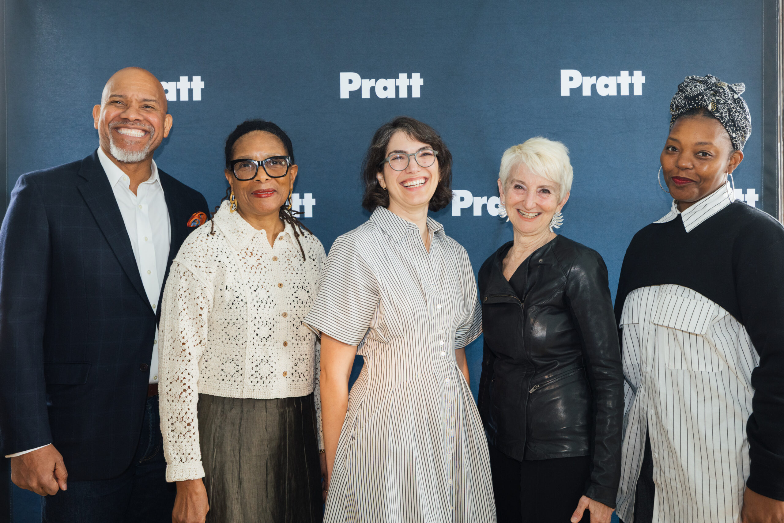 A group of five people pose for a photo in front of a blue backdrop with the word "Pratt" displayed prominently. The individuals include a man with a shaved head and beard, a woman wearing glasses and a white, lace-like top, a woman in a striped dress, a woman with short, white hair in a black leather jacket, and a woman in a black top with white stripes. They are all smiling and standing closely together.