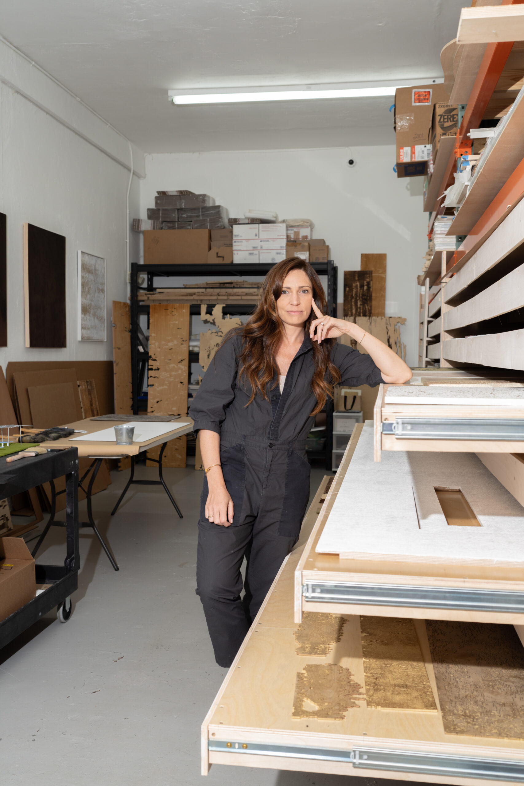 Joyce Billet stands in an art studio, leaning on a table filled with various materials. The background shows shelves with boxes and artworks. Bright lighting illuminates the room, highlighting the organized workspace.