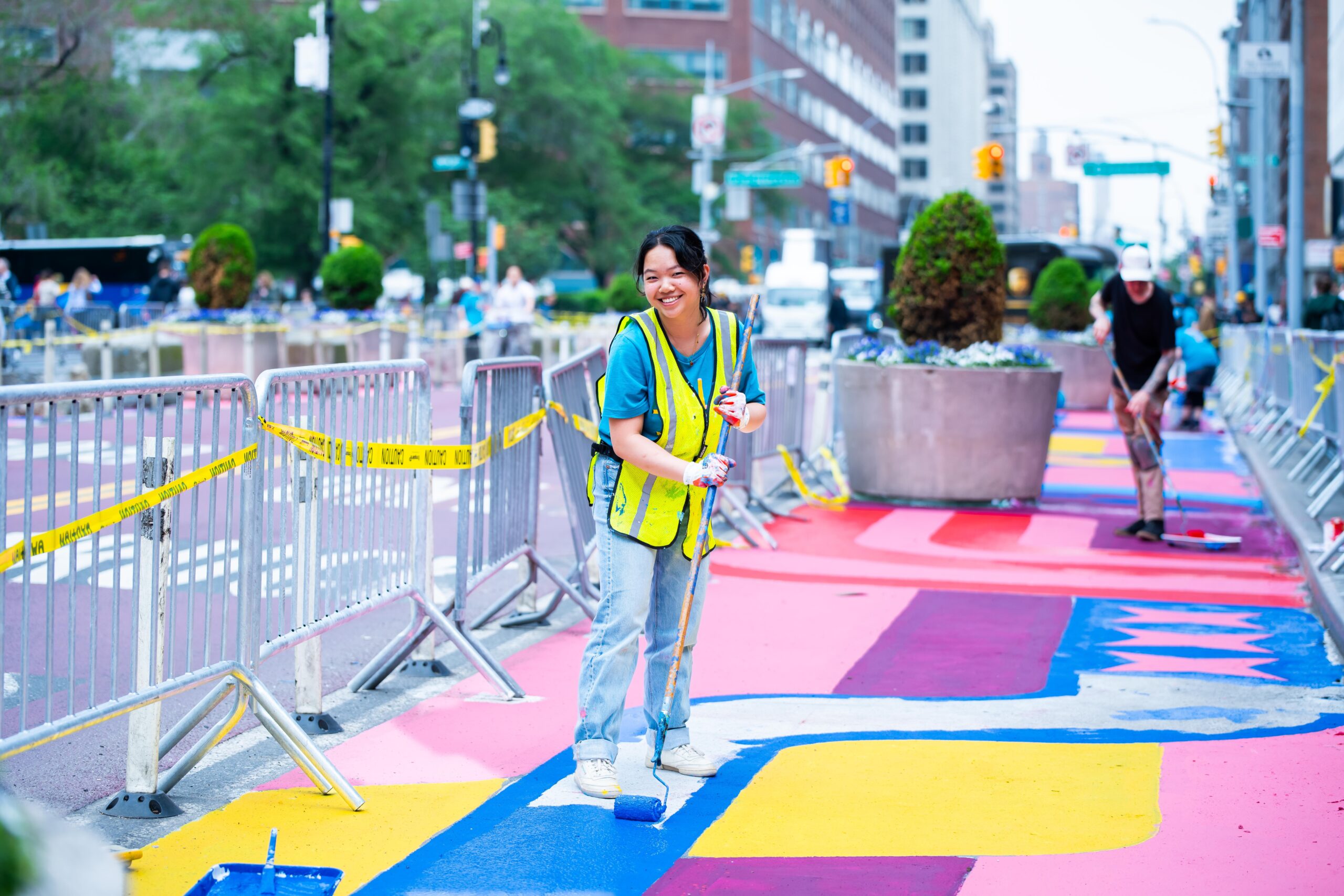 A young woman in a reflective yellow vest smiles while painting a colorful mural on a street. She holds a paint roller and stands on bright pink and blue sections of the artwork. In the background, there are other people working on the mural and construction barriers are set up along the edge of the street. Trees and urban buildings are visible in the background, with a lively street scene.
