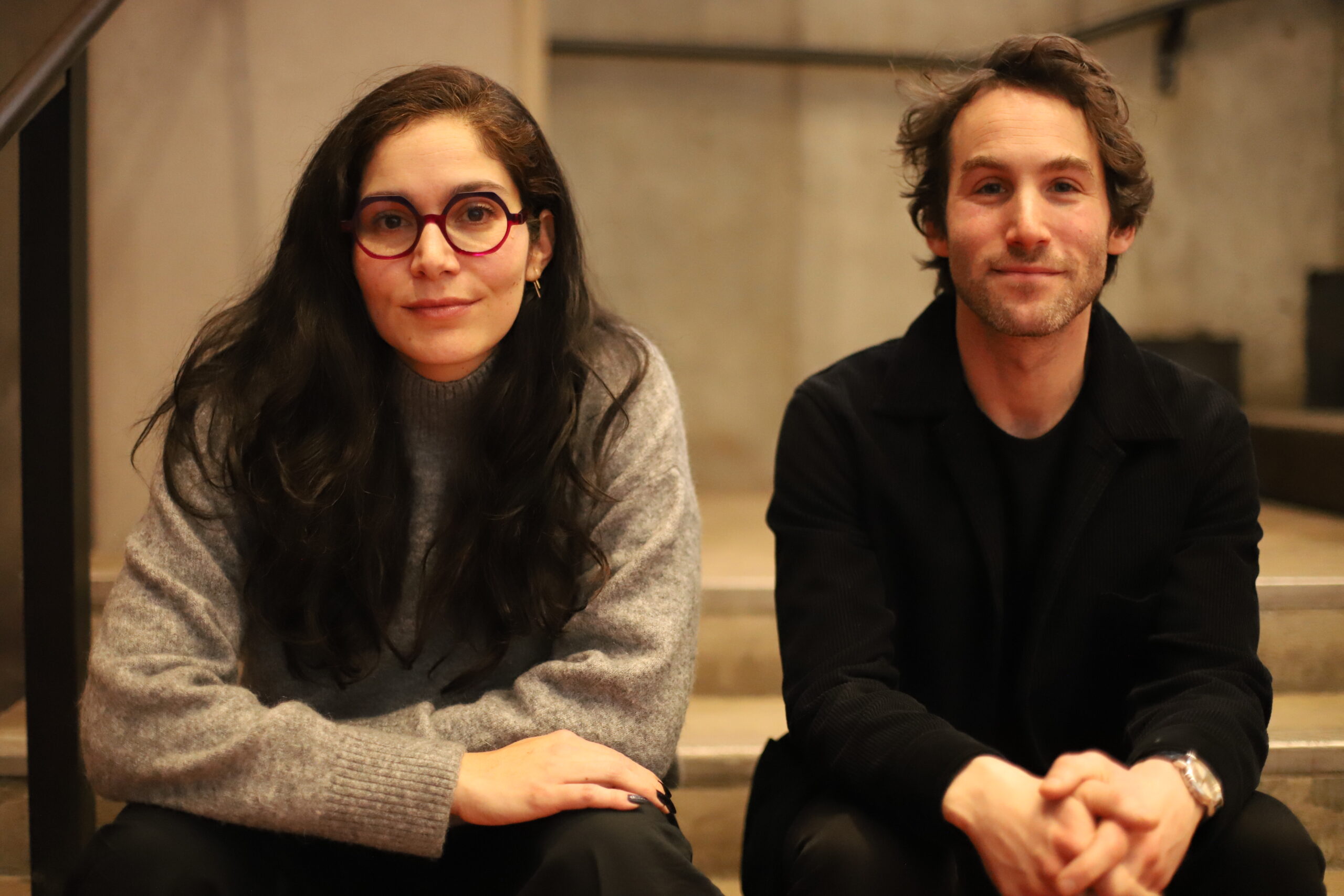 A woman with long dark hair and round, pink glasses wears a gray sweater as she sits on steps next to a man who has wavy brown hair and a light beard. He is dressed in a black jacket and smiles at the camera. The background features a minimalist, industrial-style interior with concrete walls.