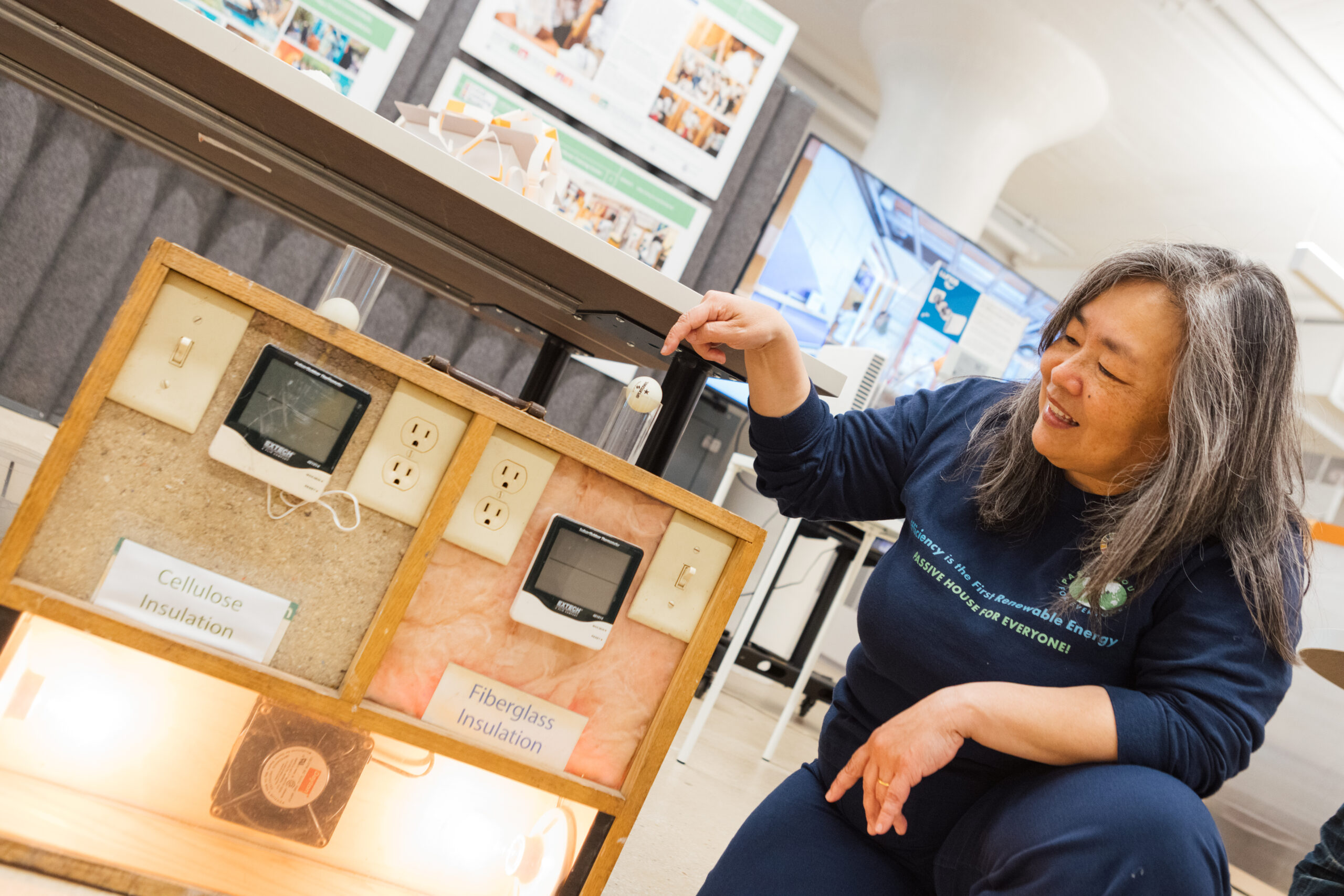 A woman is kneeling beside a display featuring two types of insulation: cellulose and fiberglass. There are two digital thermometers mounted on the display, showing different readings. The woman is pointing to a small spherical object above the insulation while smiling. In the background, there are informational posters and a monitor displaying data related to energy efficiency.