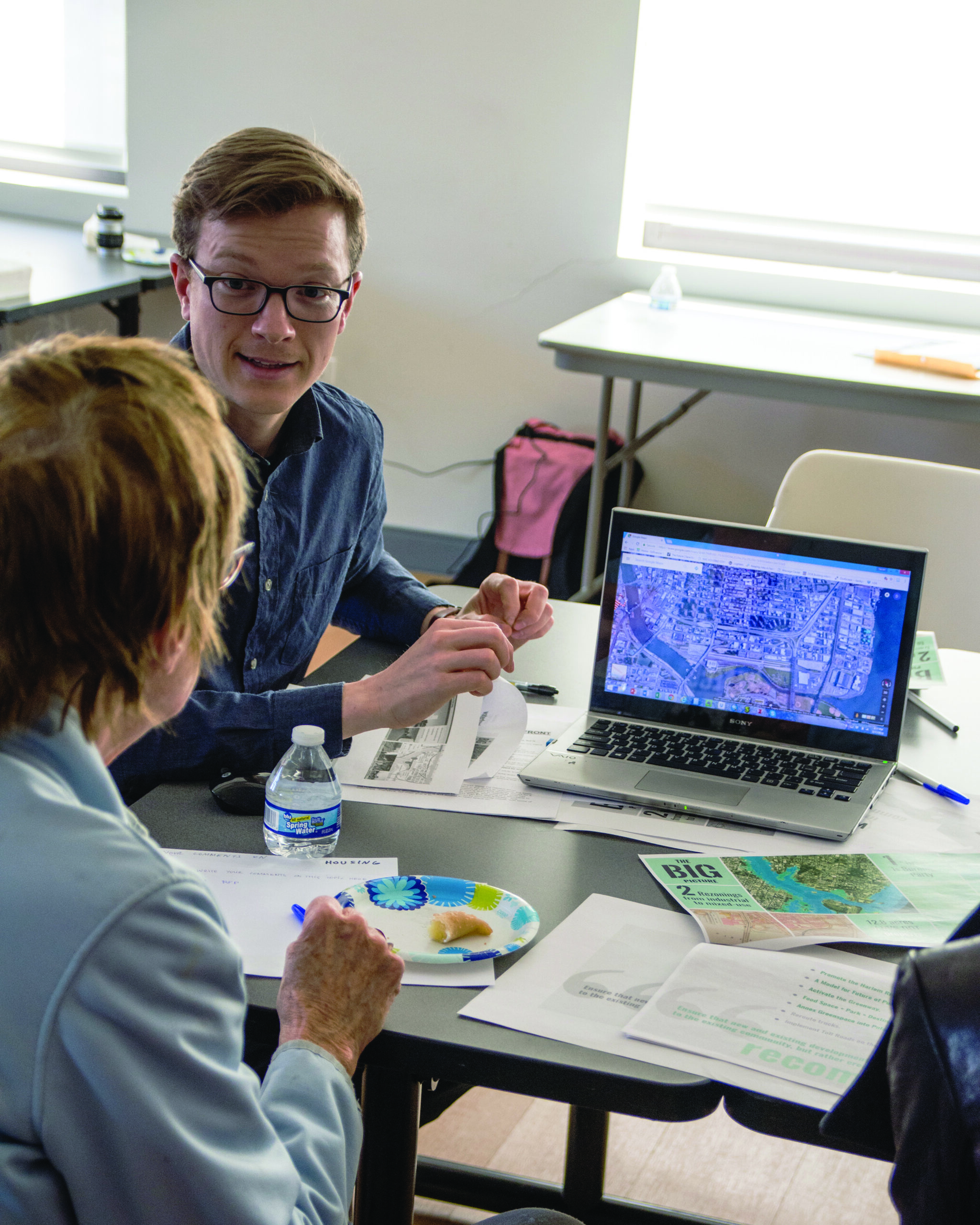 two people sit at a table with a laptop open between them. there is an image of a map on the screen. there are pieces of paper with different diagrams and drawings on them on the table.