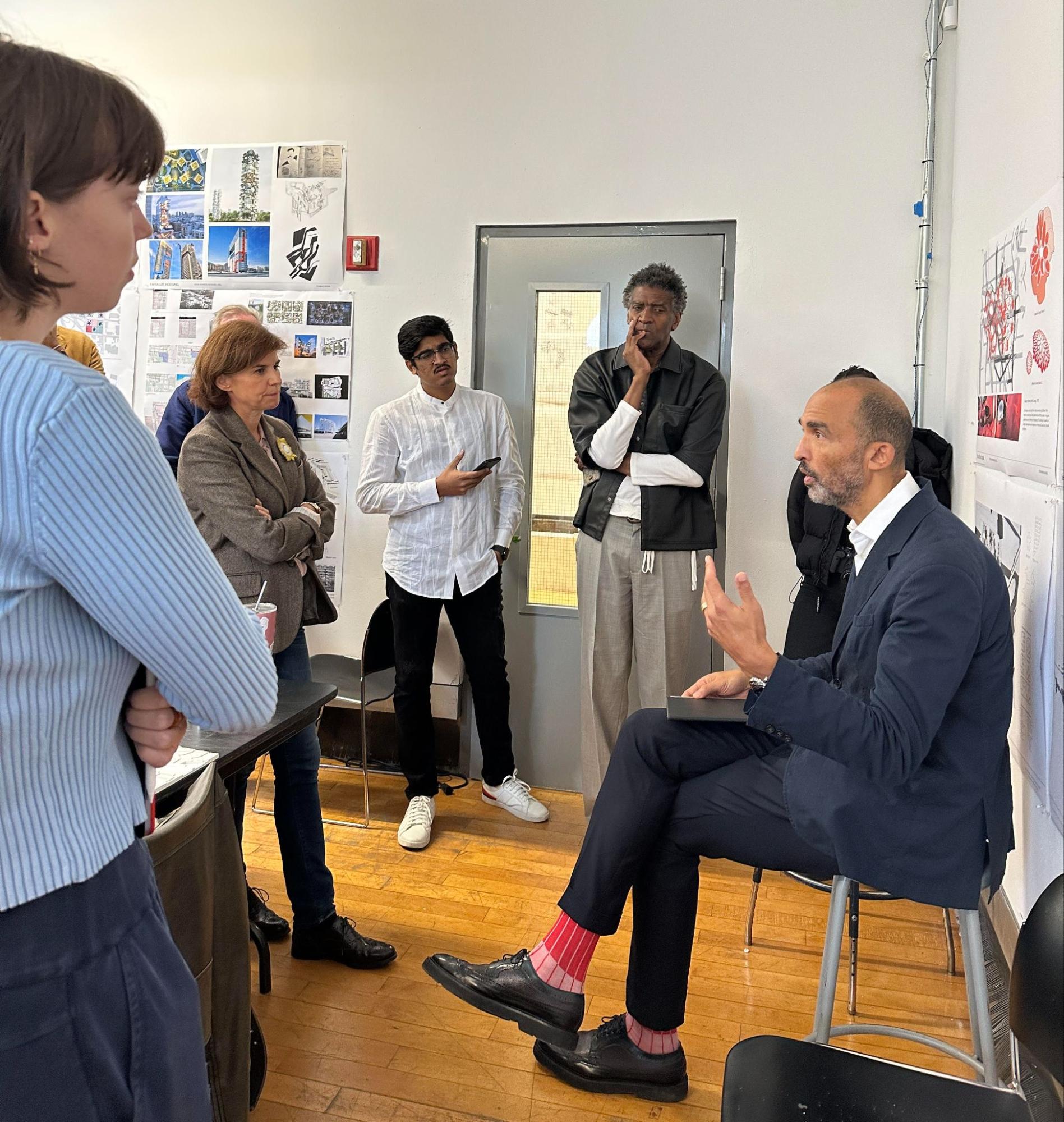 people in a classroom, standing around a man in a suit, seated. the man is speaking to the group.