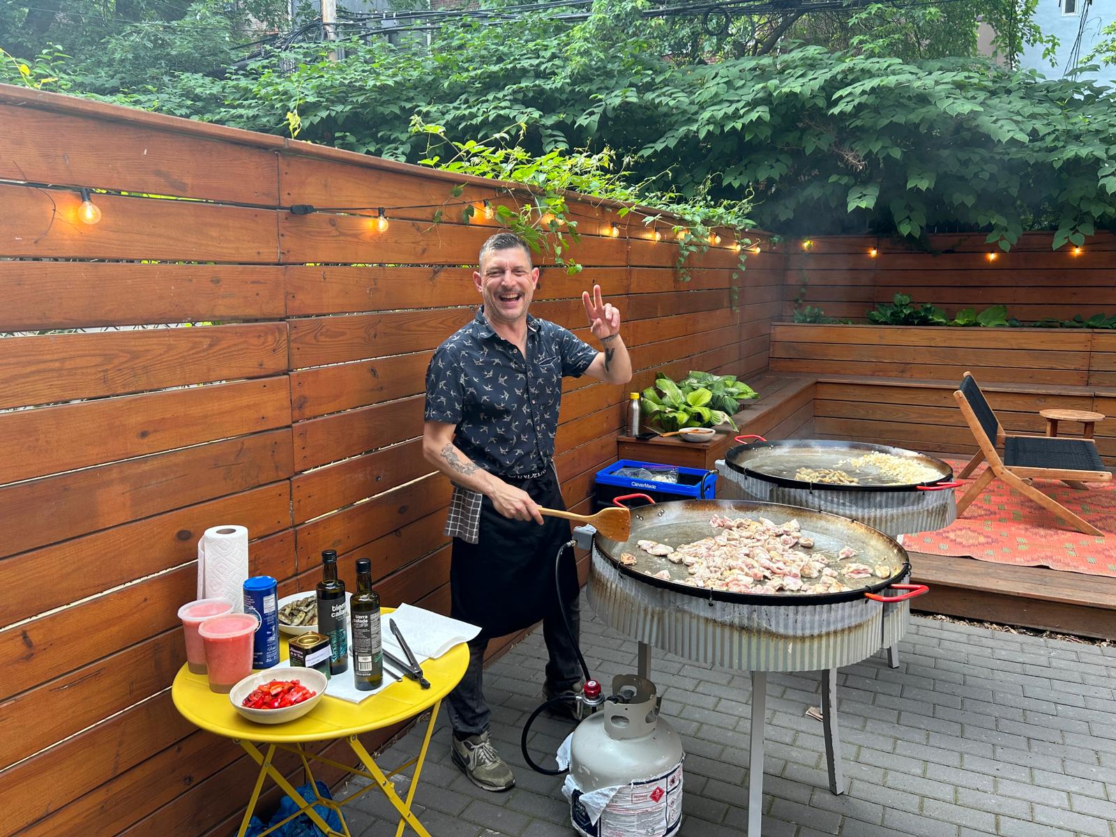 A cheerful man, wearing a patterned shirt and apron, stands next to two large pans of food cooking on propane burners in an outdoor setting. He smiles and gives a peace sign with one hand while holding a cooking utensil with the other. The cooking area is adorned with string lights and surrounded by lush green plants and wooden fencing, creating a warm and inviting ambiance. Various ingredients and cooking tools are neatly arranged on a small yellow table beside him.