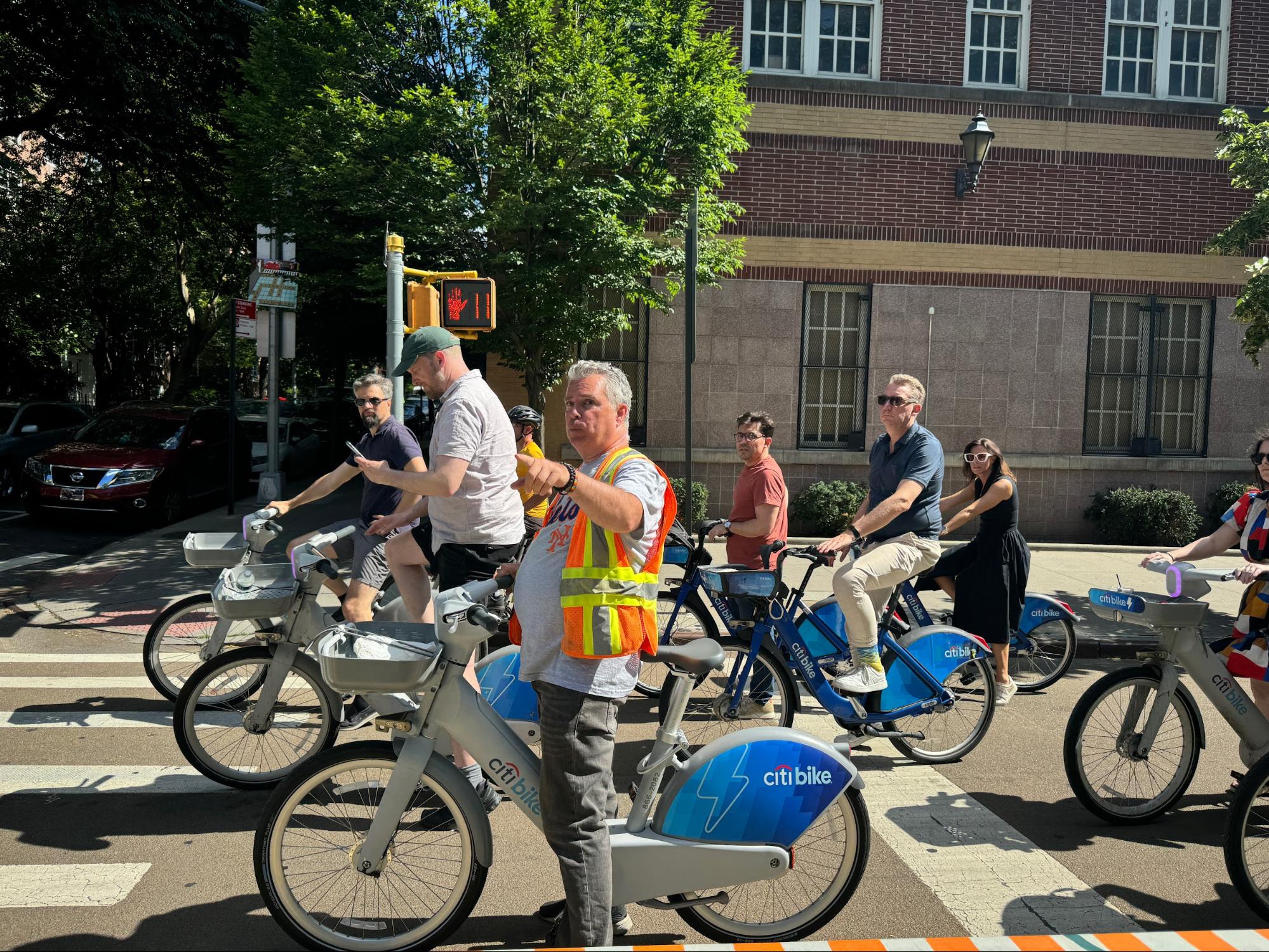 A group of people on Citi Bikes, stopped at a street intersection, with a mix of casual and professional attire. One person in a reflective vest is pointing ahead, possibly giving directions. The setting is urban with trees and buildings in the background, and the weather appears sunny and pleasant.