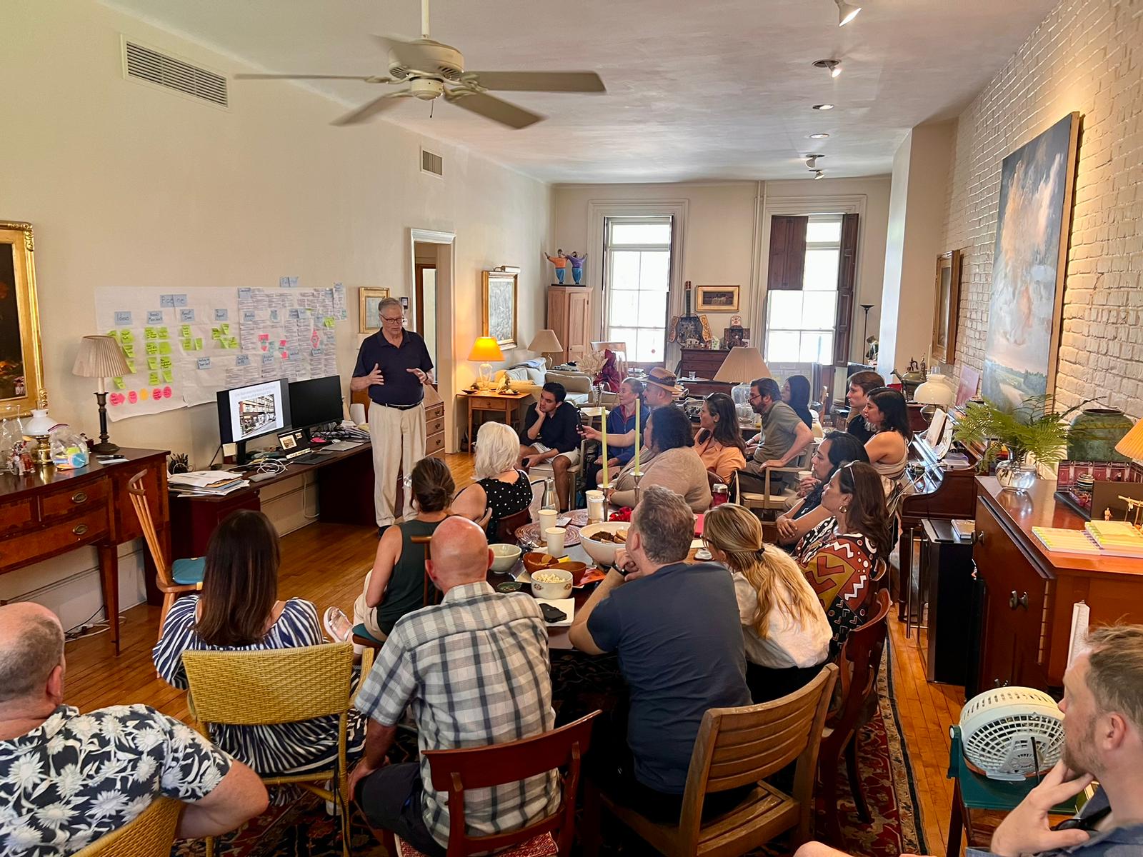 A group of people attentively listens to a speaker in a cozy, well-lit room. The speaker stands near a wall covered with colorful sticky notes and large sheets of paper filled with handwritten notes and diagrams. The attendees, seated in a semi-circle, are engaged in the discussion, creating a collaborative and interactive atmosphere. The room is decorated with wooden furniture and artwork, adding a homely feel to the professional setting.