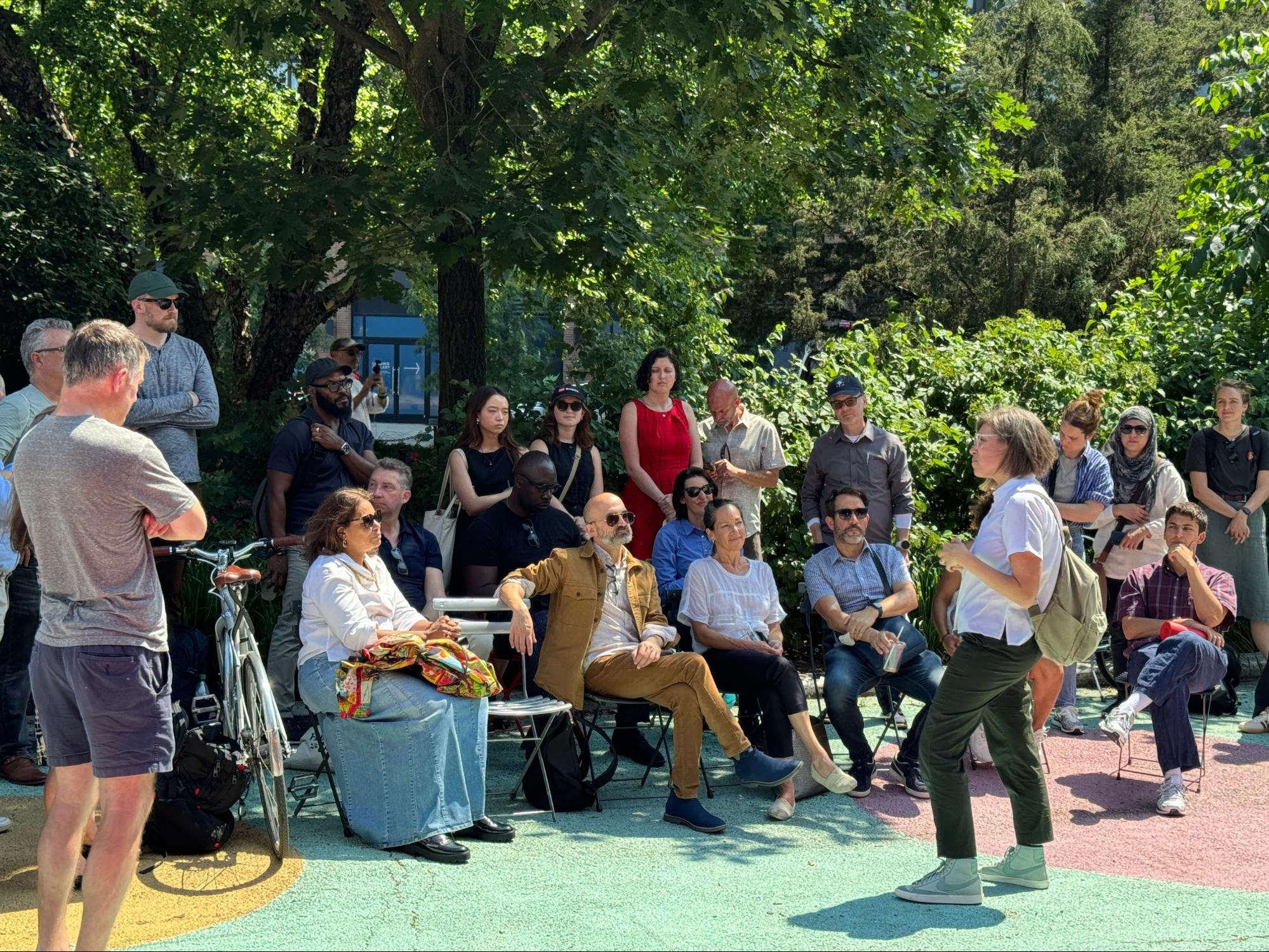 A woman with short hair and glasses, wearing a white shirt and dark pants, is standing and speaking to a group of people seated and standing around her in an outdoor setting. The audience is diverse, with some sitting on chairs and others standing, surrounded by lush greenery and sunlight filtering through the trees. The mood is informal and engaged, indicating an outdoor meeting or educational talk.
