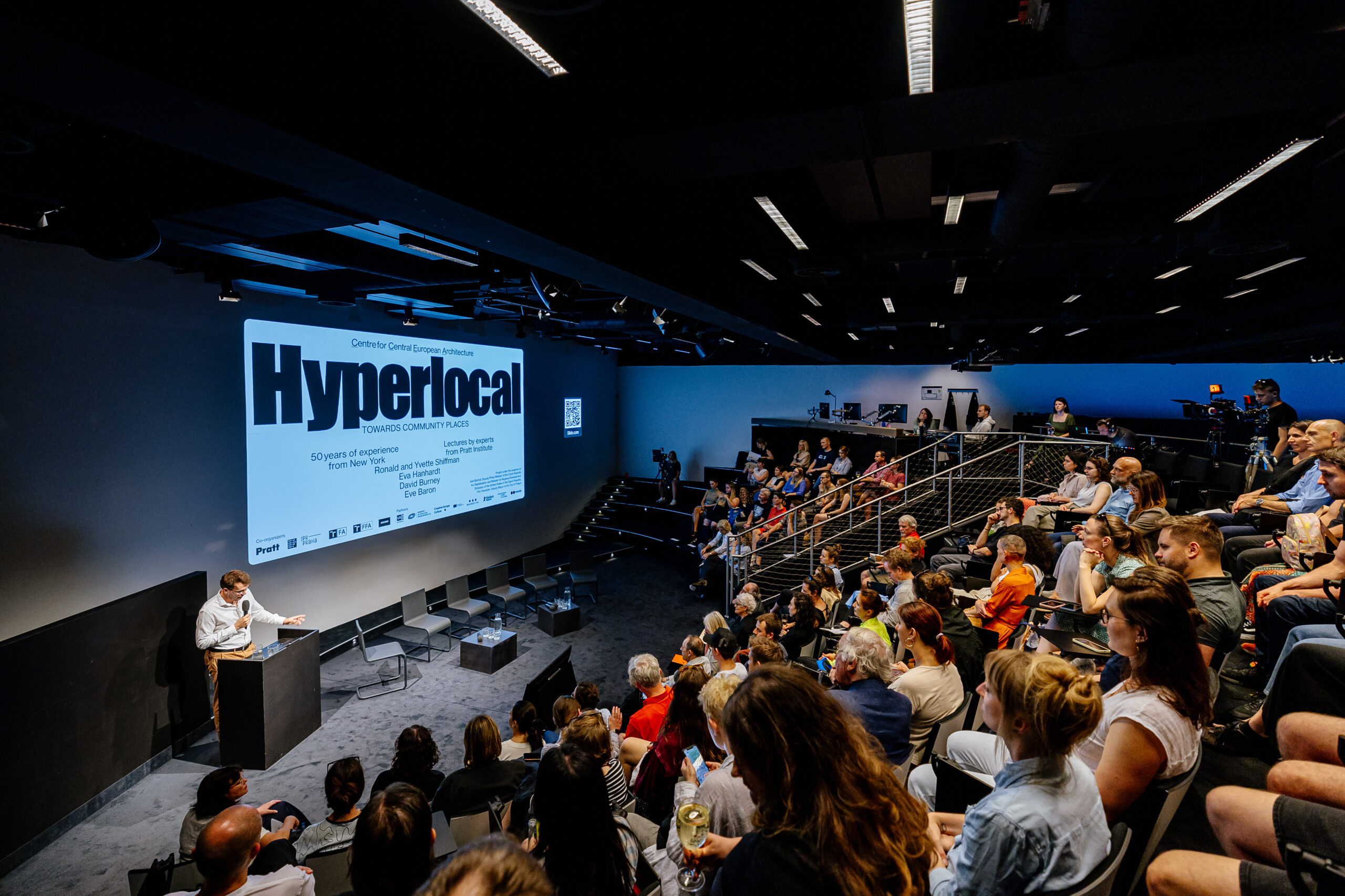 A lecture event in a modern, theater-style auditorium. The presentation screen displays a slide titled "Hyperlocal" with details about the lecture series. The audience is seated and attentively watching a speaker at a podium.