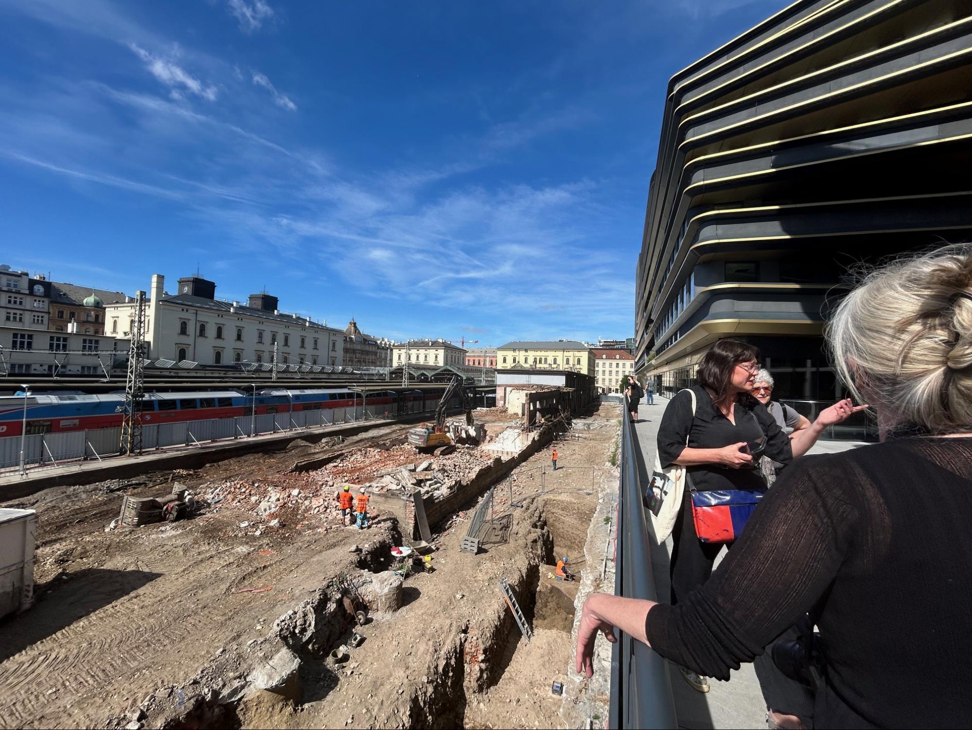 A group of people standing on a bridge overlooking a train station construction site, with bright blue skies and buildings in the background.